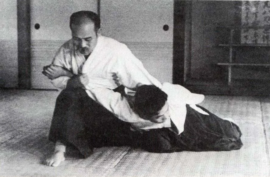 Two martial artists practicing a grappling technique on a tatami mat in a traditional dojo, with shoji screens and wooden display cabinets in the background.