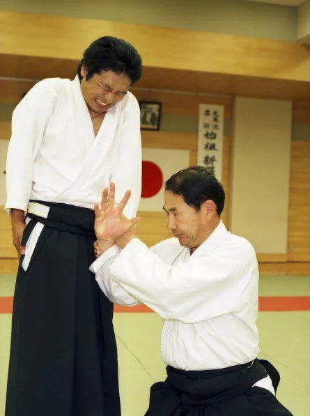 Two martial artists practicing a technique in a dojo, with Japanese calligraphy on the wall behind them.