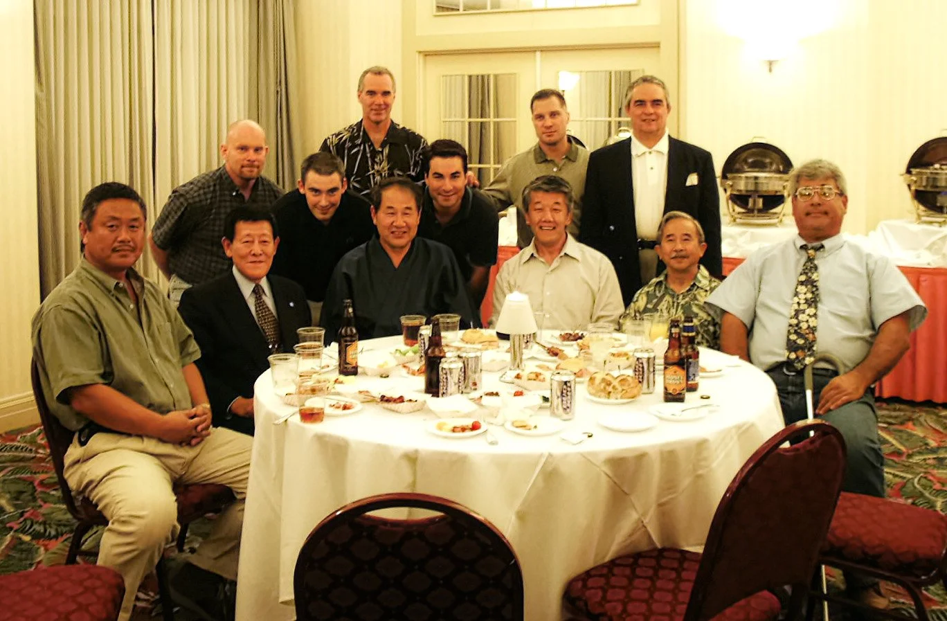 A group of fifteen men gathered around a banquet table at a formal event in a hotel dining room with buffet stations in the background. The table has food and drinks, some men are seated, others standing behind them.
