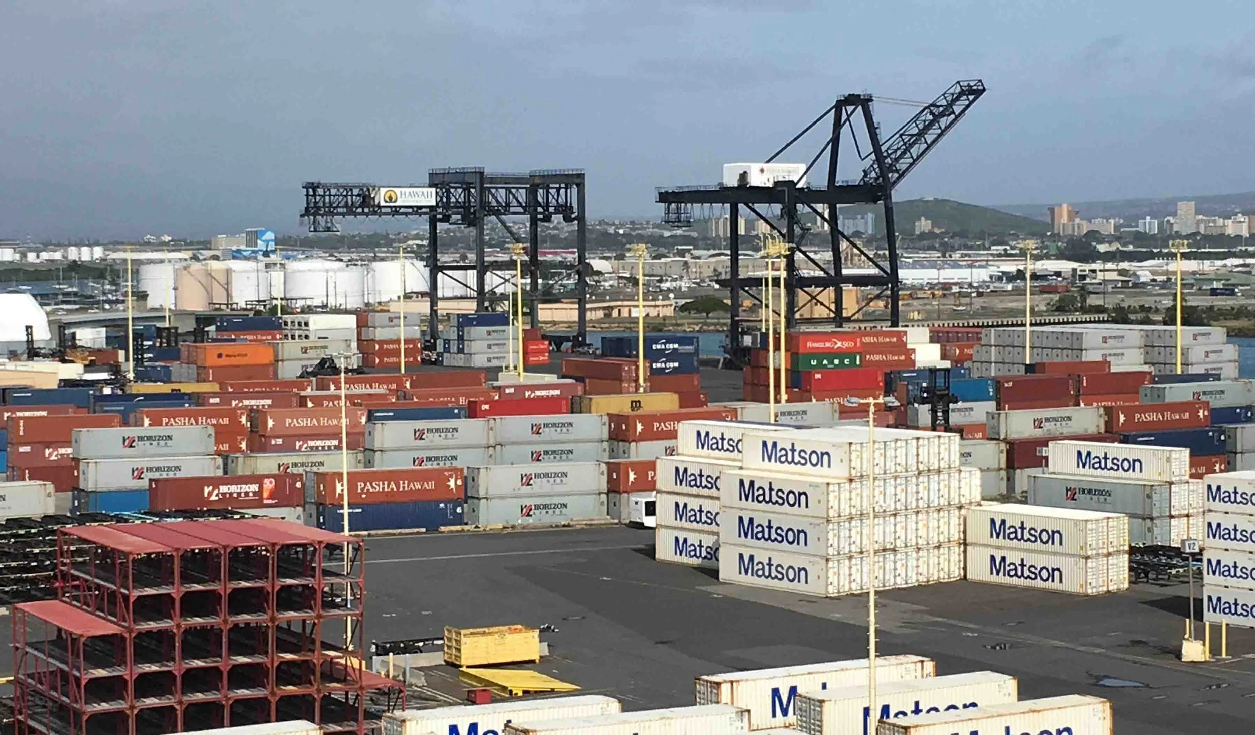 Stacks of colorful shipping containers and large industrial gantry cranes crowd an active commercial shipping terminal in Honolulu Harbor beneath an overcast sky.