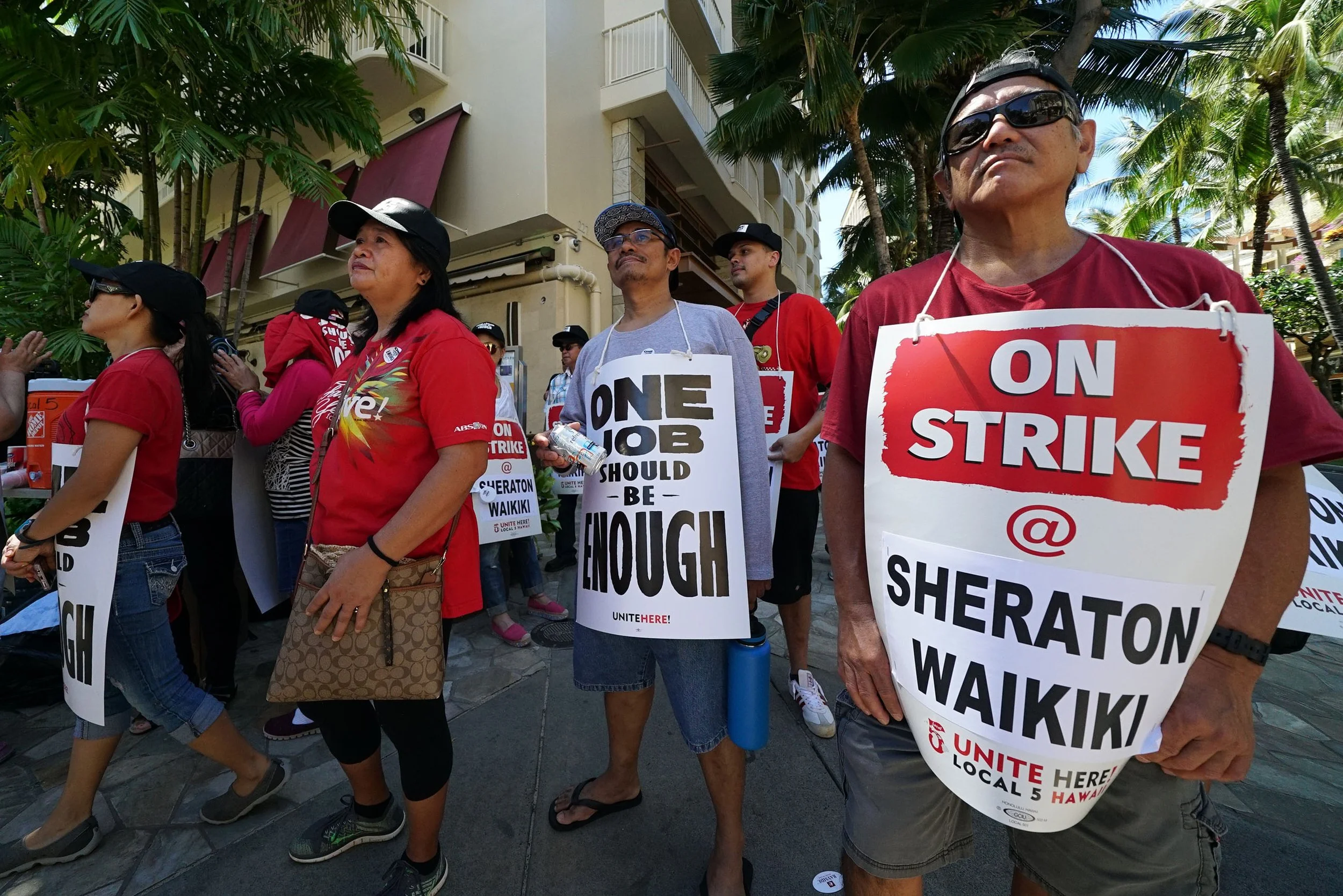Striking hotel workers in Waikiki wear picket signs reading "One Job Should Be Enough" and "On Strike @ Sheraton Waikiki" while gathered along a palm-lined sidewalk.