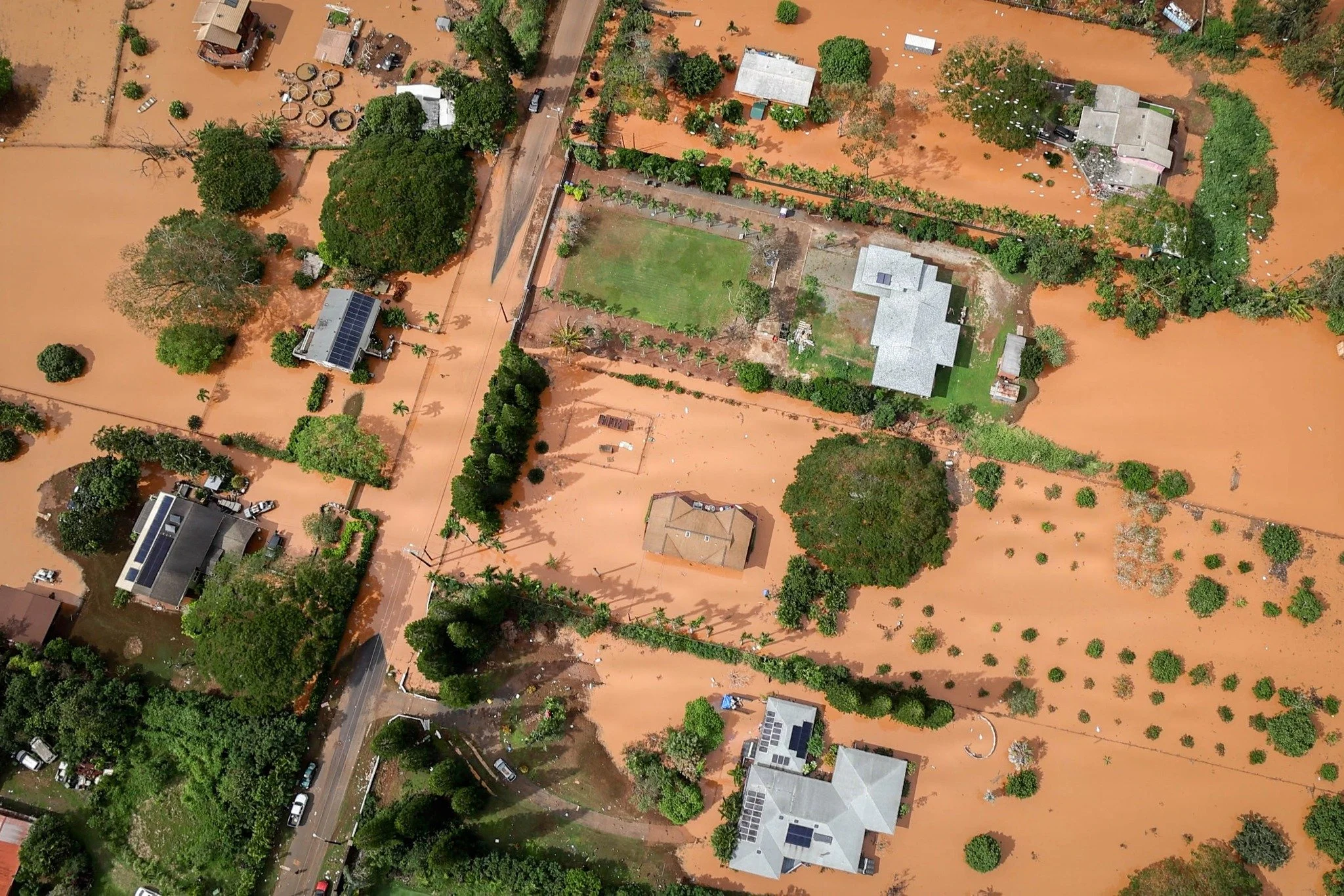 Aerial view of a residential neighborhood in Waialua, O'ahu, submerged in thick, reddish-brown floodwaters, with homes, yards, and roads surrounded by muddy inundation.