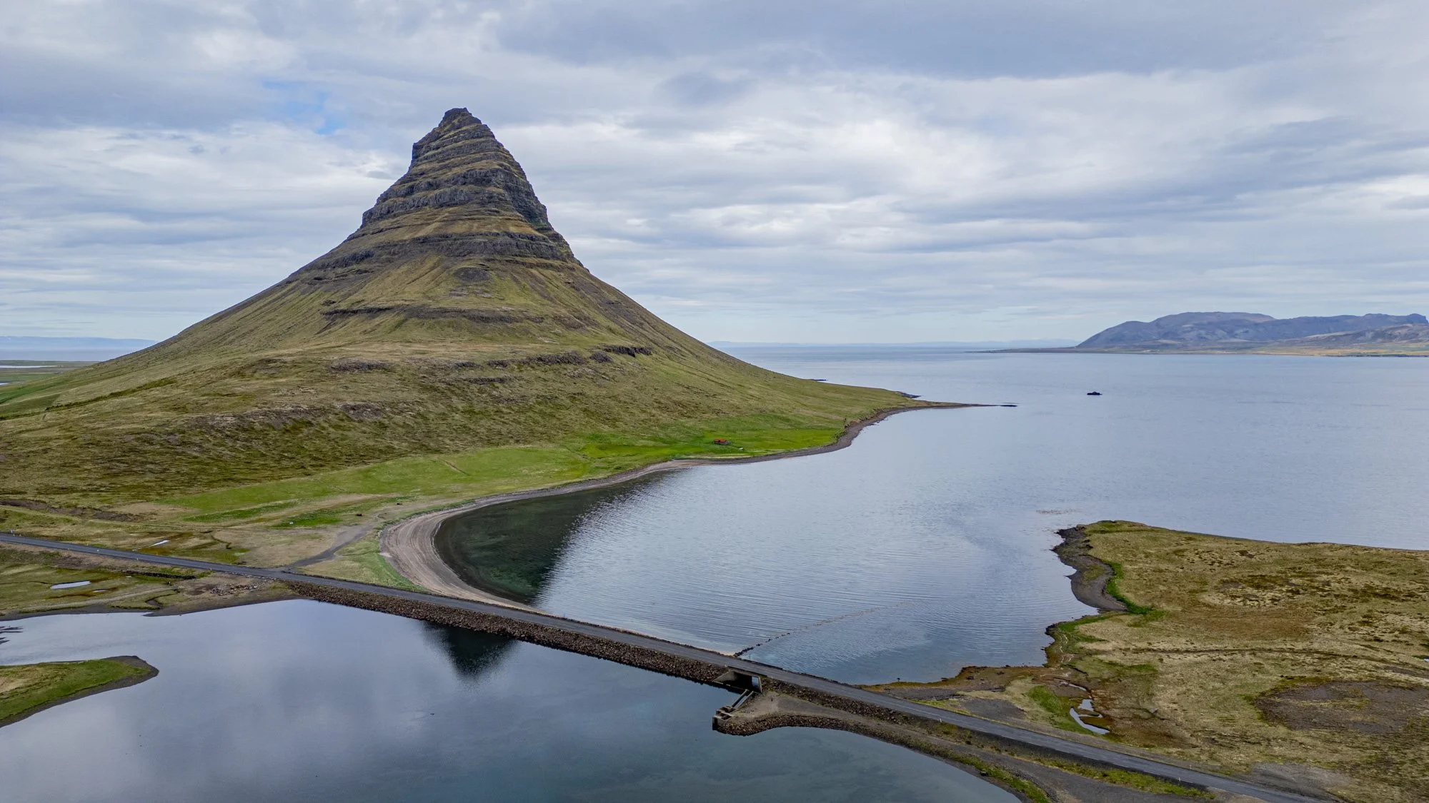 A distinctive cone-shaped mountain in Kirkjufell, Iceland, rises above a calm coastal bay crossed by a narrow causeway road under an overcast sky.