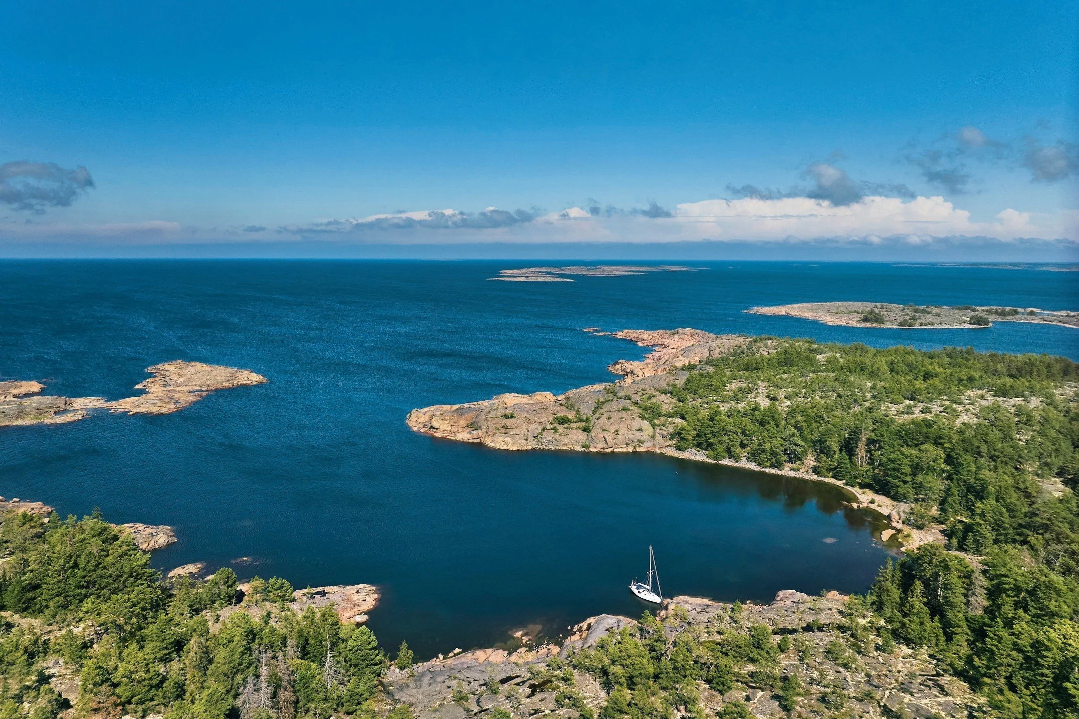 Aerial view of a sailboat anchored in a secluded, deep-blue rocky cove bordered by pine trees and island outcrops under a vast clear sky.