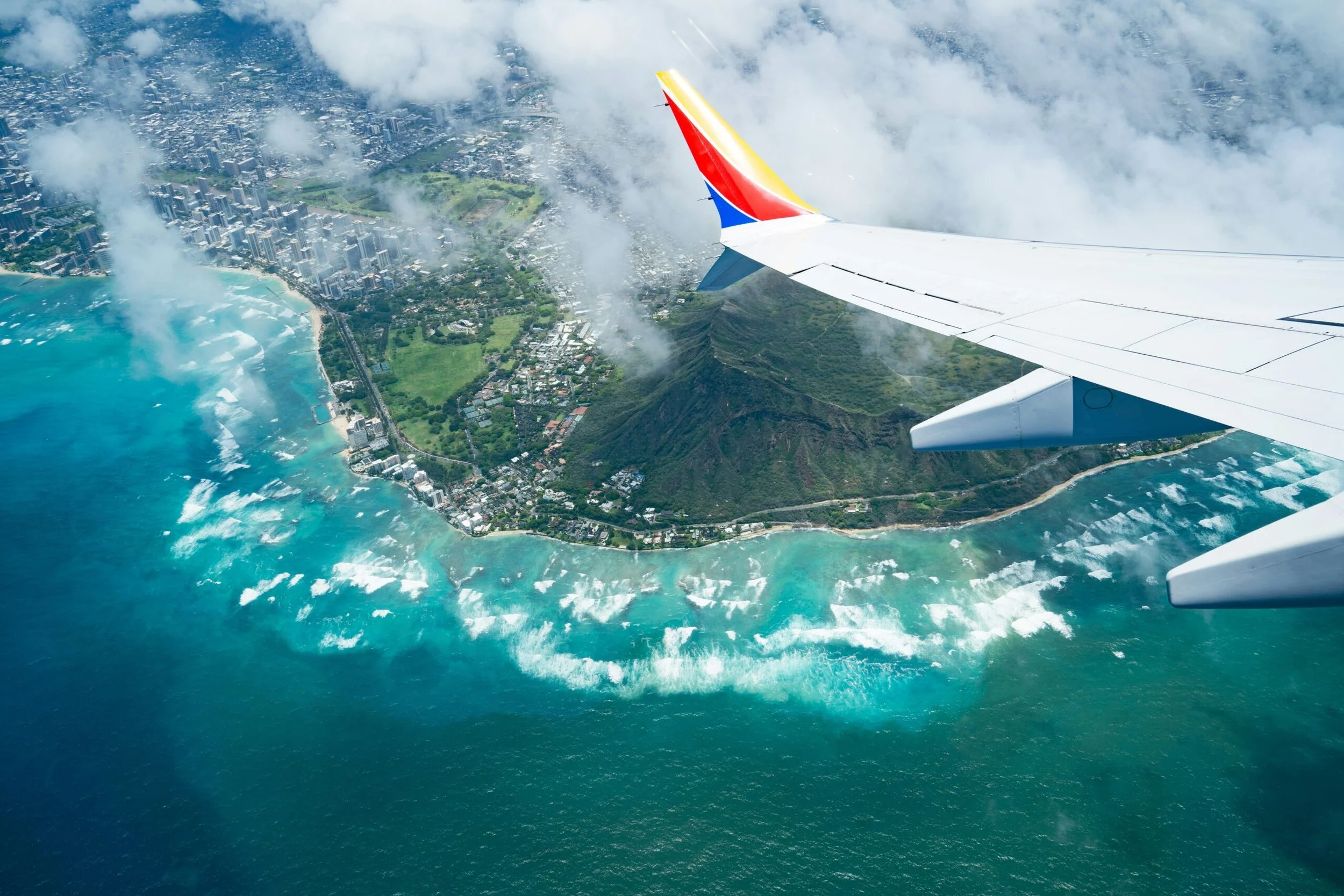 Aerial view from an airplane wing looking down at Diamond Head crater, Waikiki’s high-rises, and turquoise ocean reefs framed by scattered white clouds.