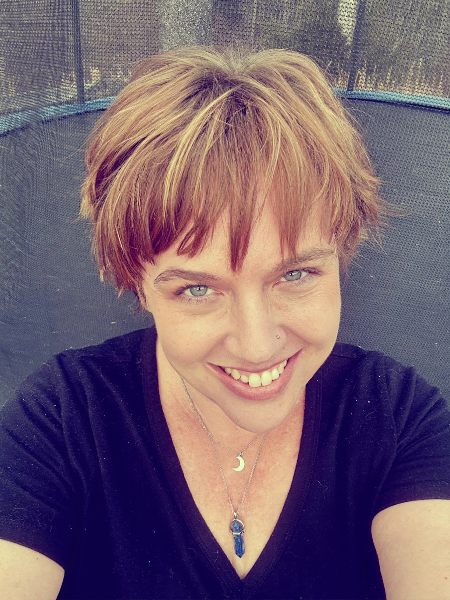 A smiling woman with short, reddish-brown hair and light-colored eyes, wearing a black shirt and layered necklaces with moon and crystal pendants, taking a selfie outdoors with a trampoline in the background.
