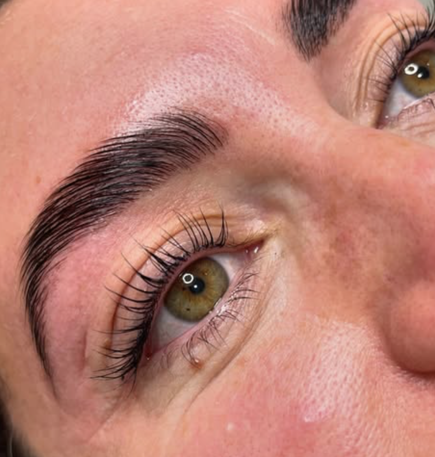 Close-up of a person's eye with well-groomed eyebrows, long curled eyelashes, and visible hair follicles on the eyelid. The person's skin appears smooth and slightly flushed.
