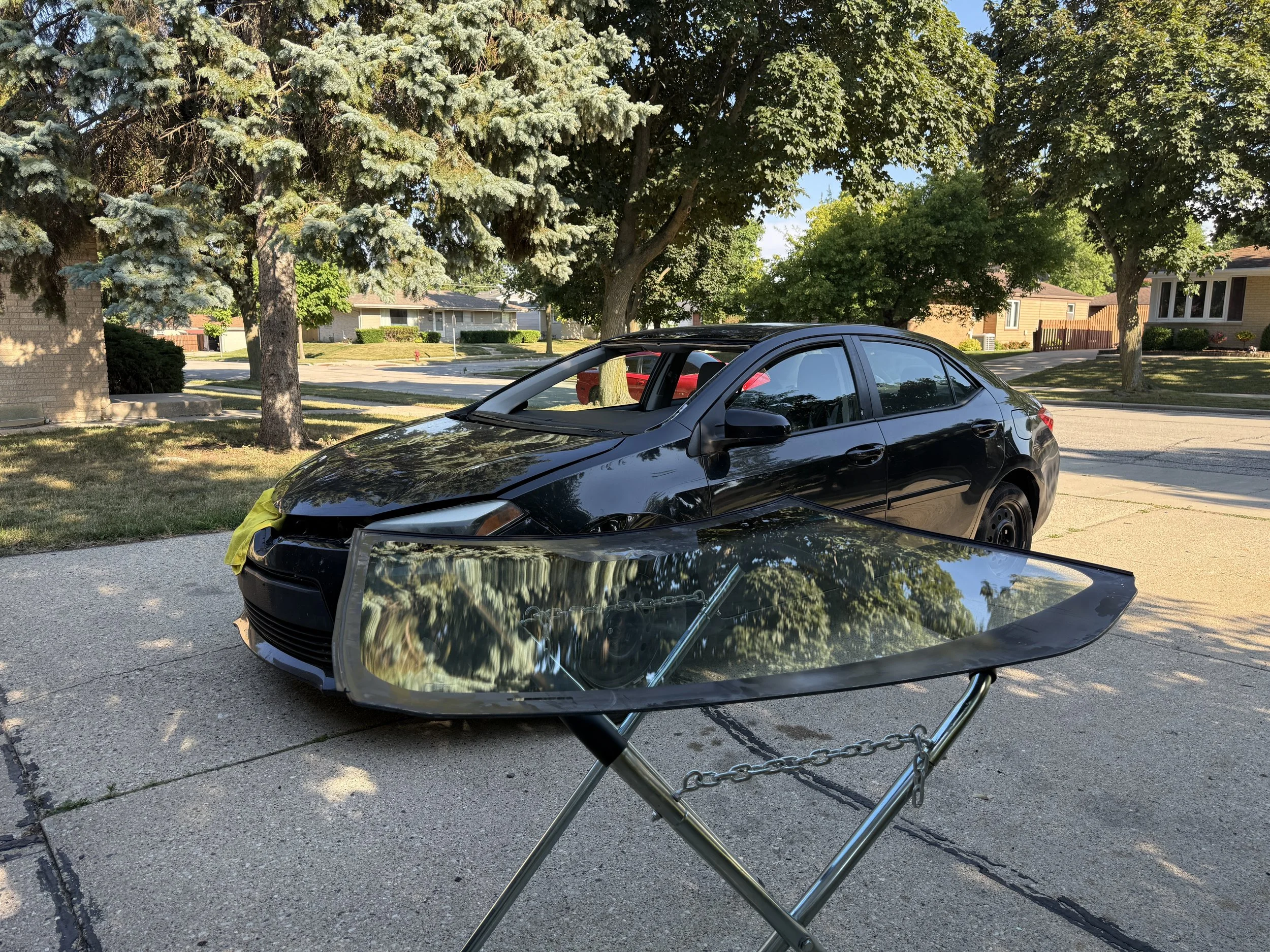 A black car with a damaged front hood parked on the street, with a small lounge chair visible in the foreground and trees and houses in the background.