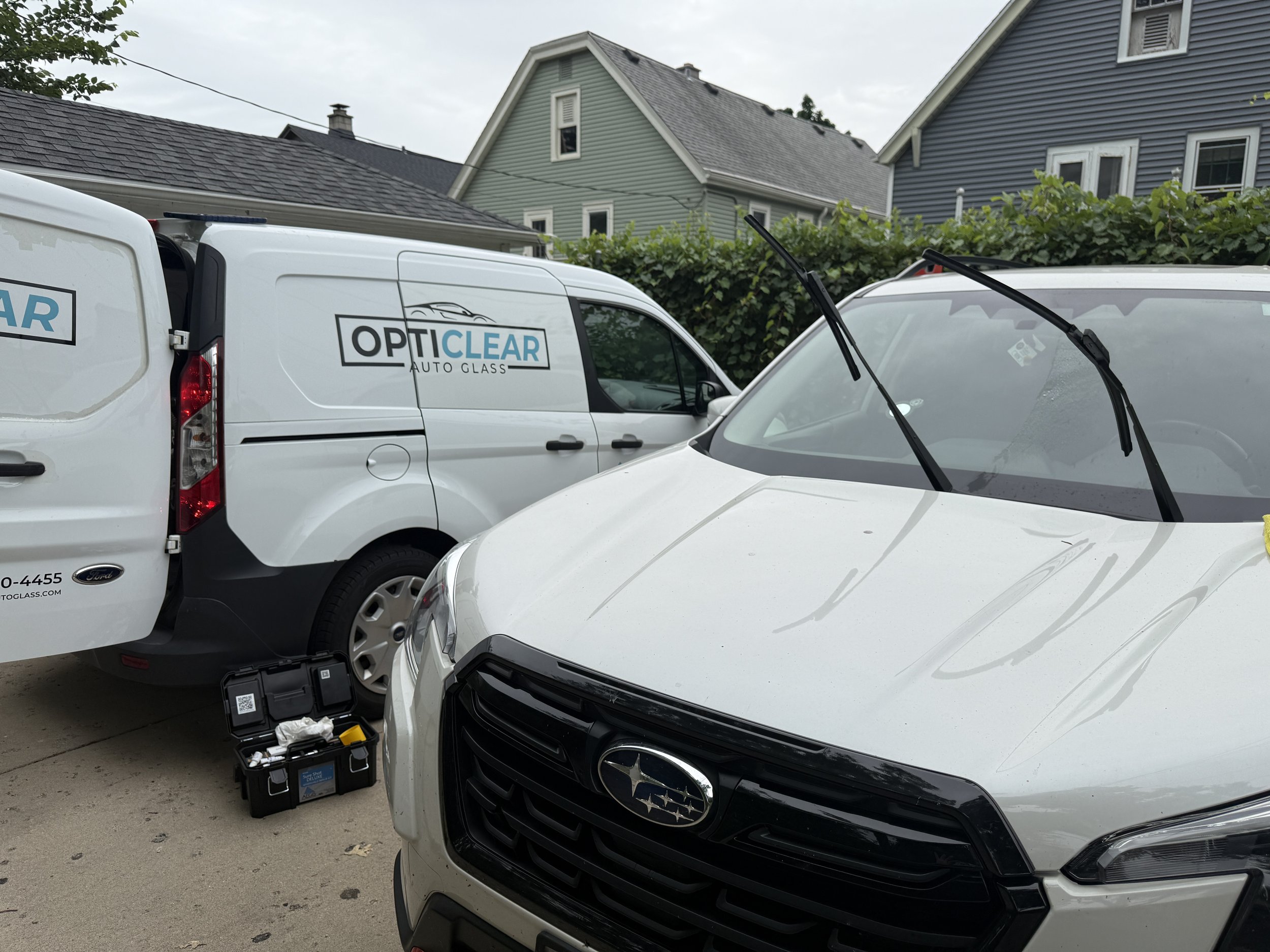 White Subaru car parked in a driveway with a black grille and hood, with maintenance tools on the ground in front. Two white vans with 'OptiClear Auto Glass' logos parked nearby. Residential houses with gray and green siding and a hedge in the background.