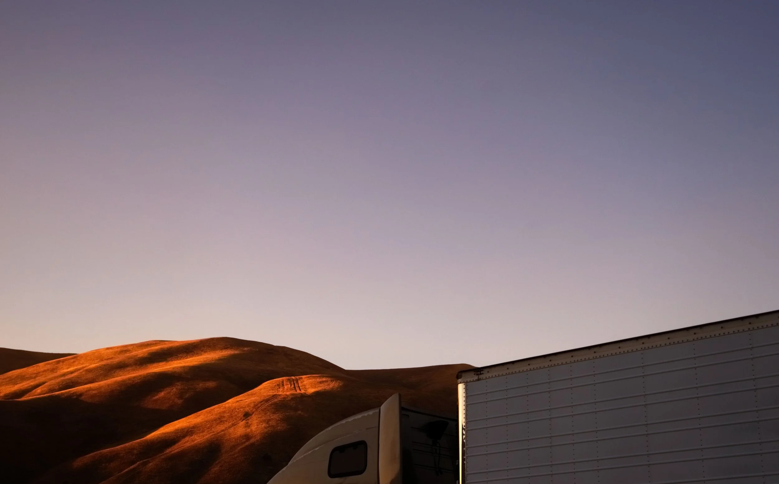 A semi truck in shadow against burnt brown hills and a violet sunset sky. Surreal fine art landscape photography.