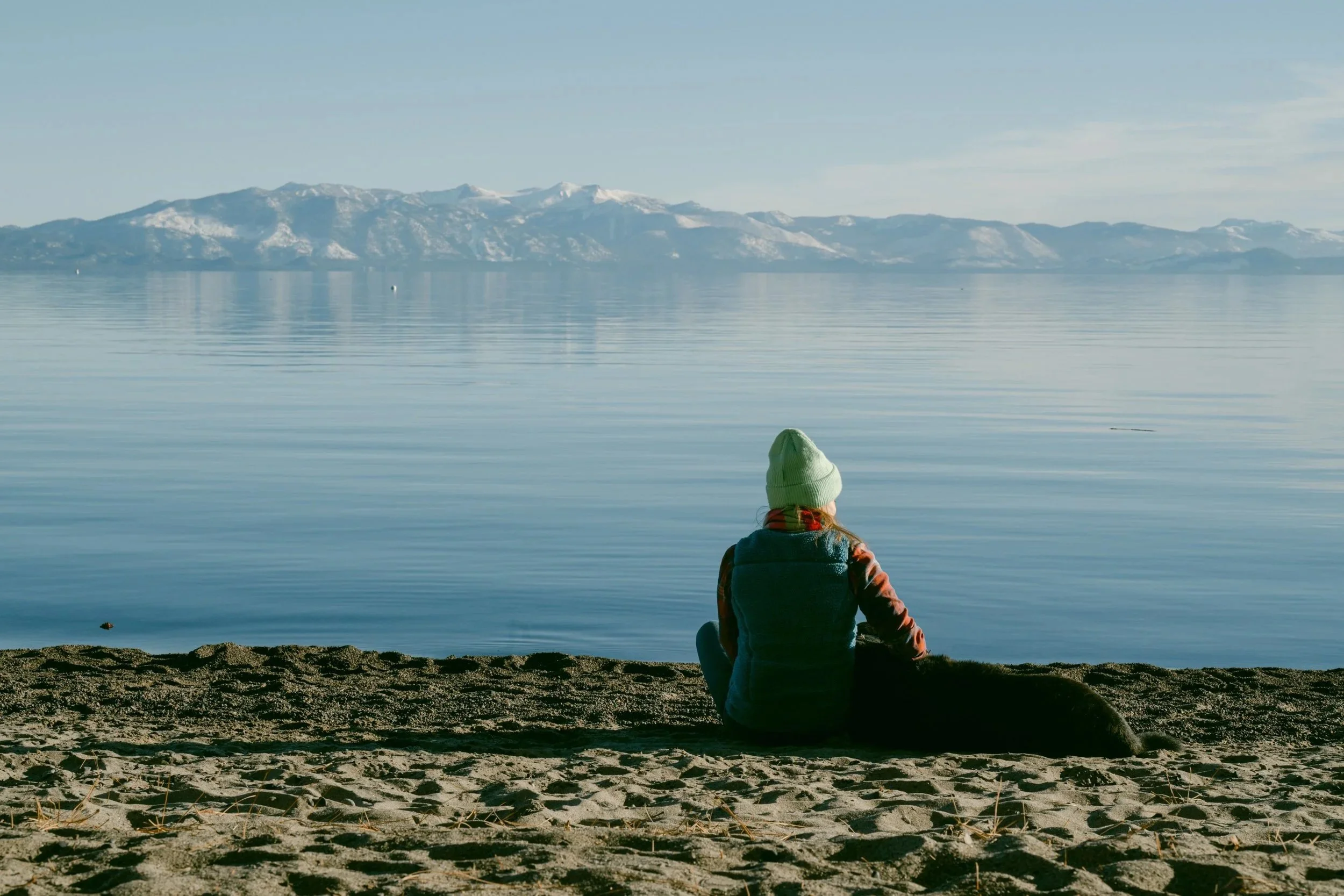 Person sitting on a sandy beach beside a black dog, looking out at a calm lake and mountains in Lake Tahoe
