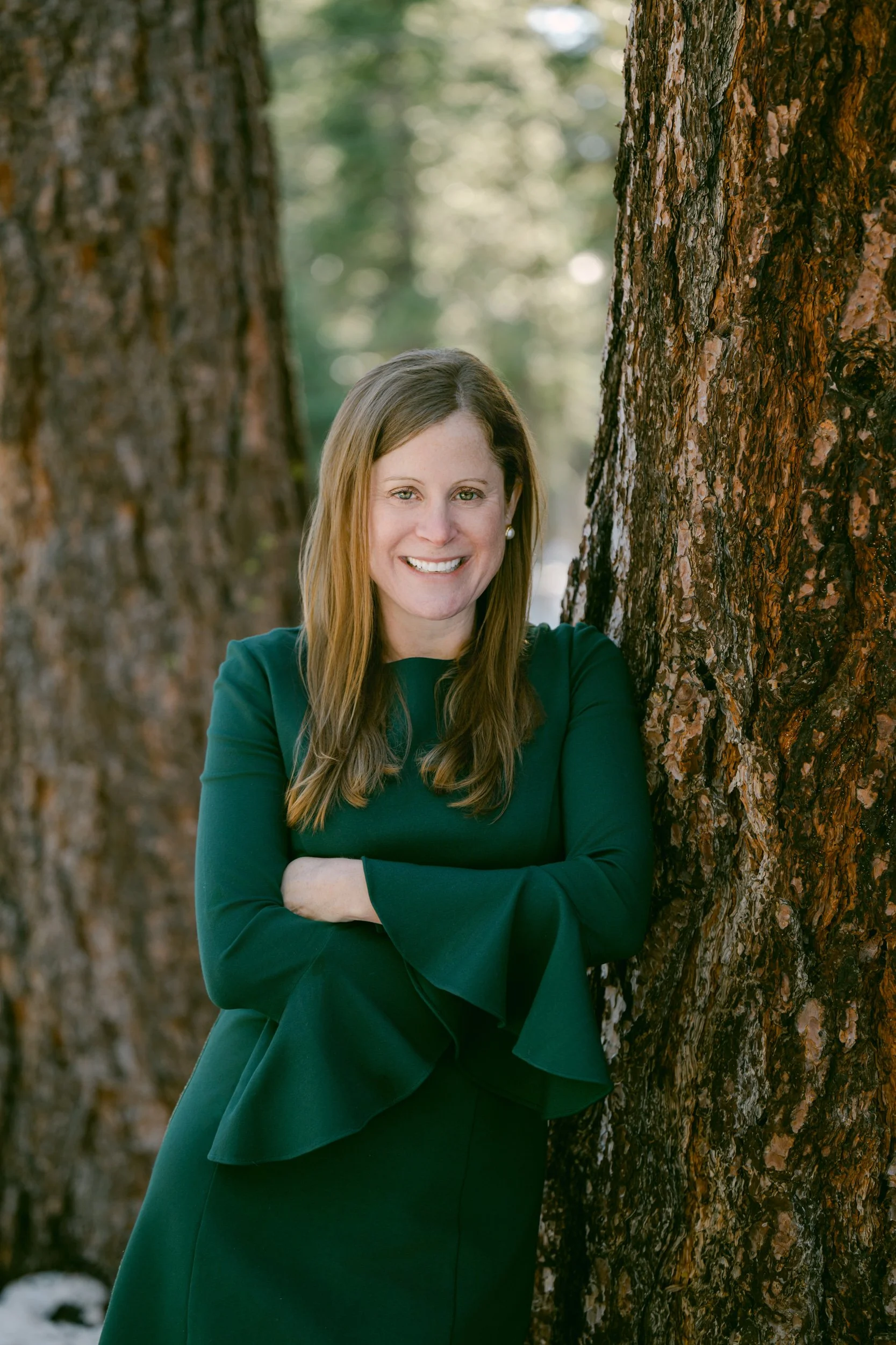 Woman Dr. Christina Harding in a dark green dress smiling beside a large pine tree outdoors