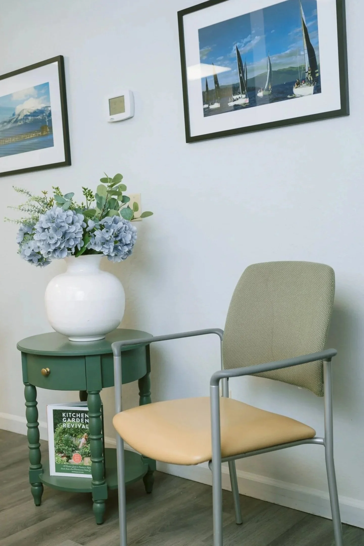 Light-filled office corner with a sage guest chair, green side table, white vase of blue hydrangeas, and framed artwork