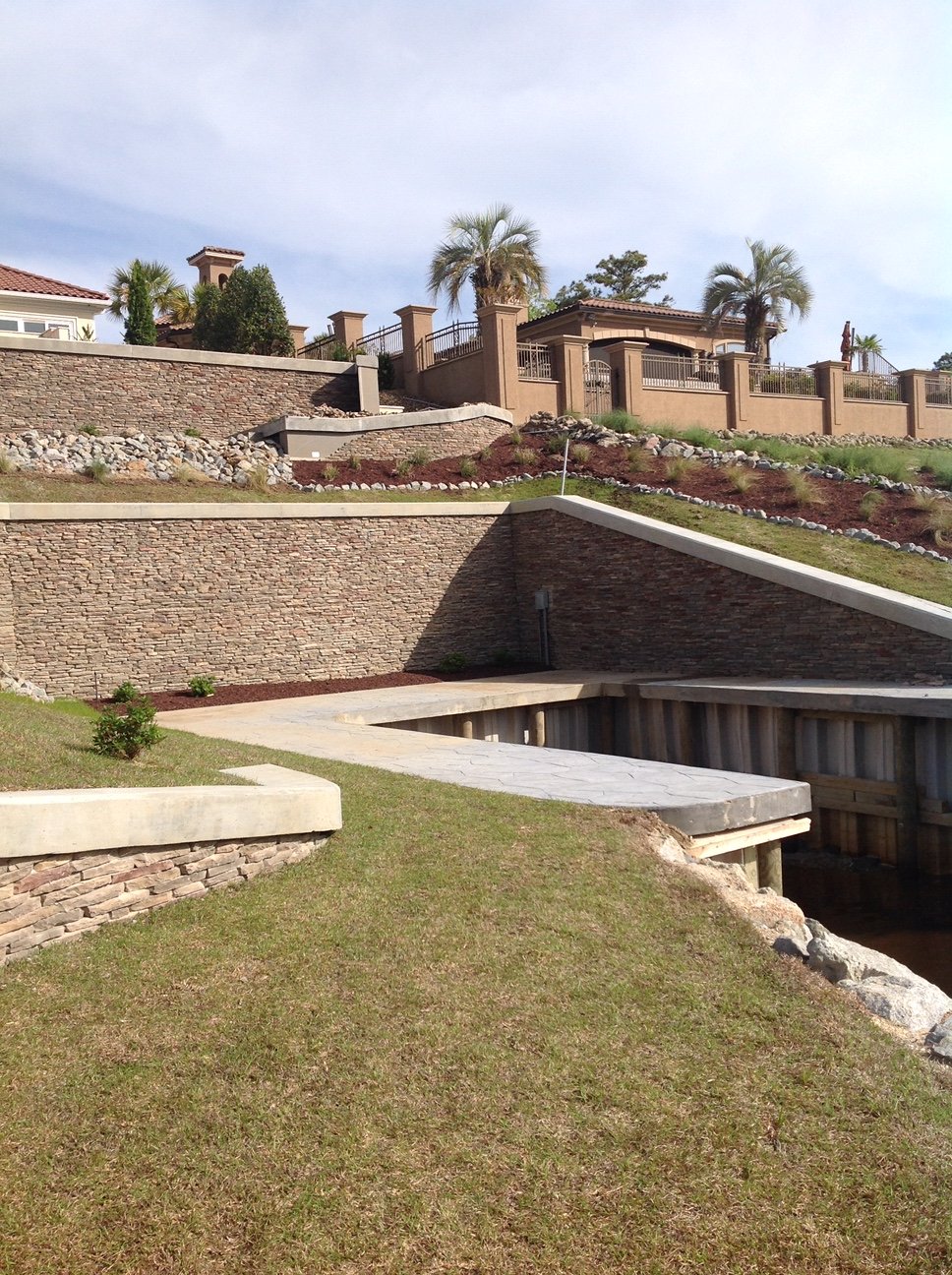 A landscaped yard with multiple levels and retaining walls made of stone and concrete, featuring palm trees, shrubs, and a house in the background.