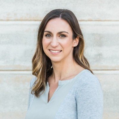A smiling woman with long wavy brown hair, wearing a light blue top, standing outdoors in front of a light-colored stone wall.