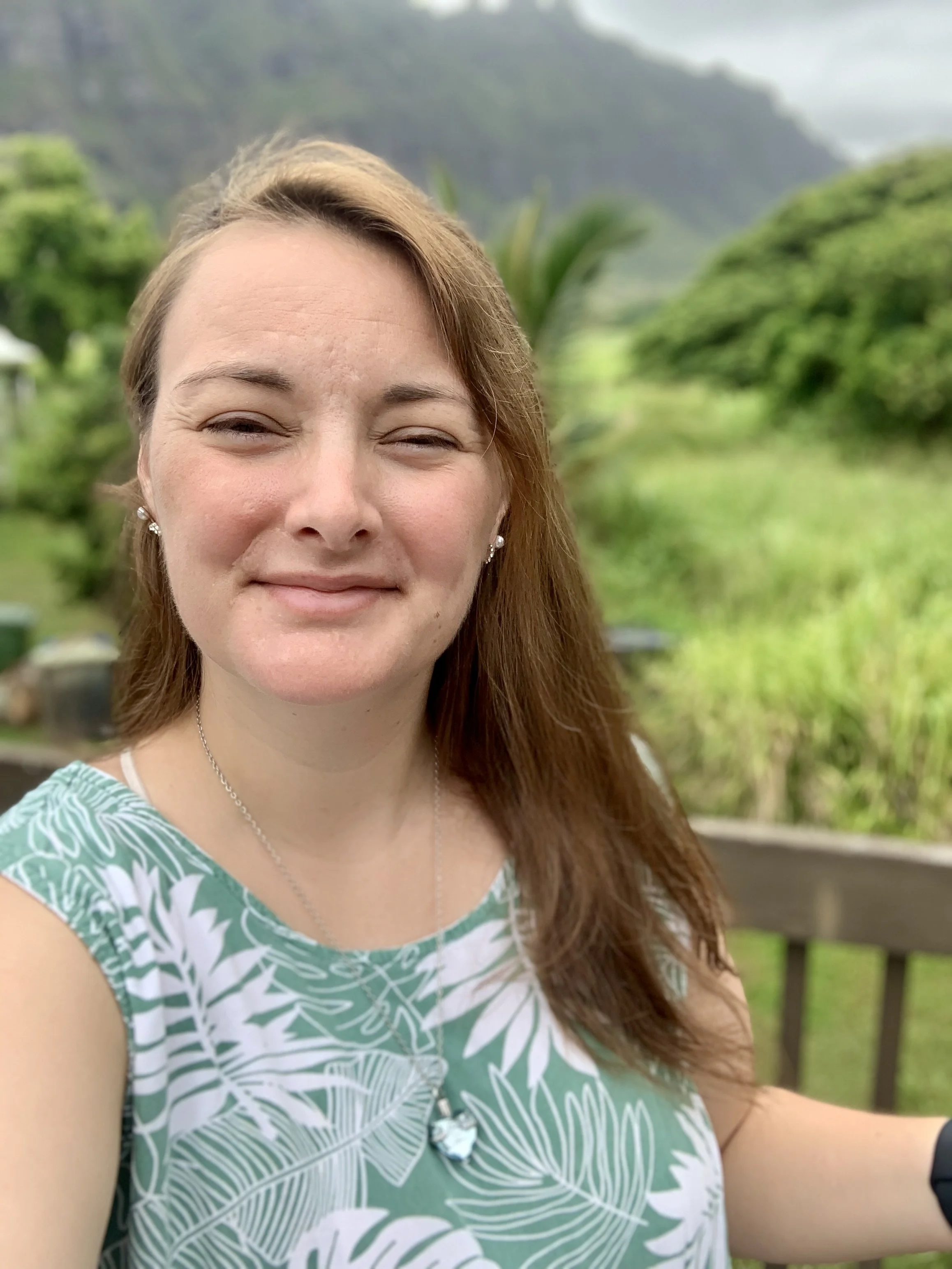 A woman with brown hair taking a selfie outdoors in a lush green, mountainous area, wearing a sleeveless green and white patterned top and silver jewelry.