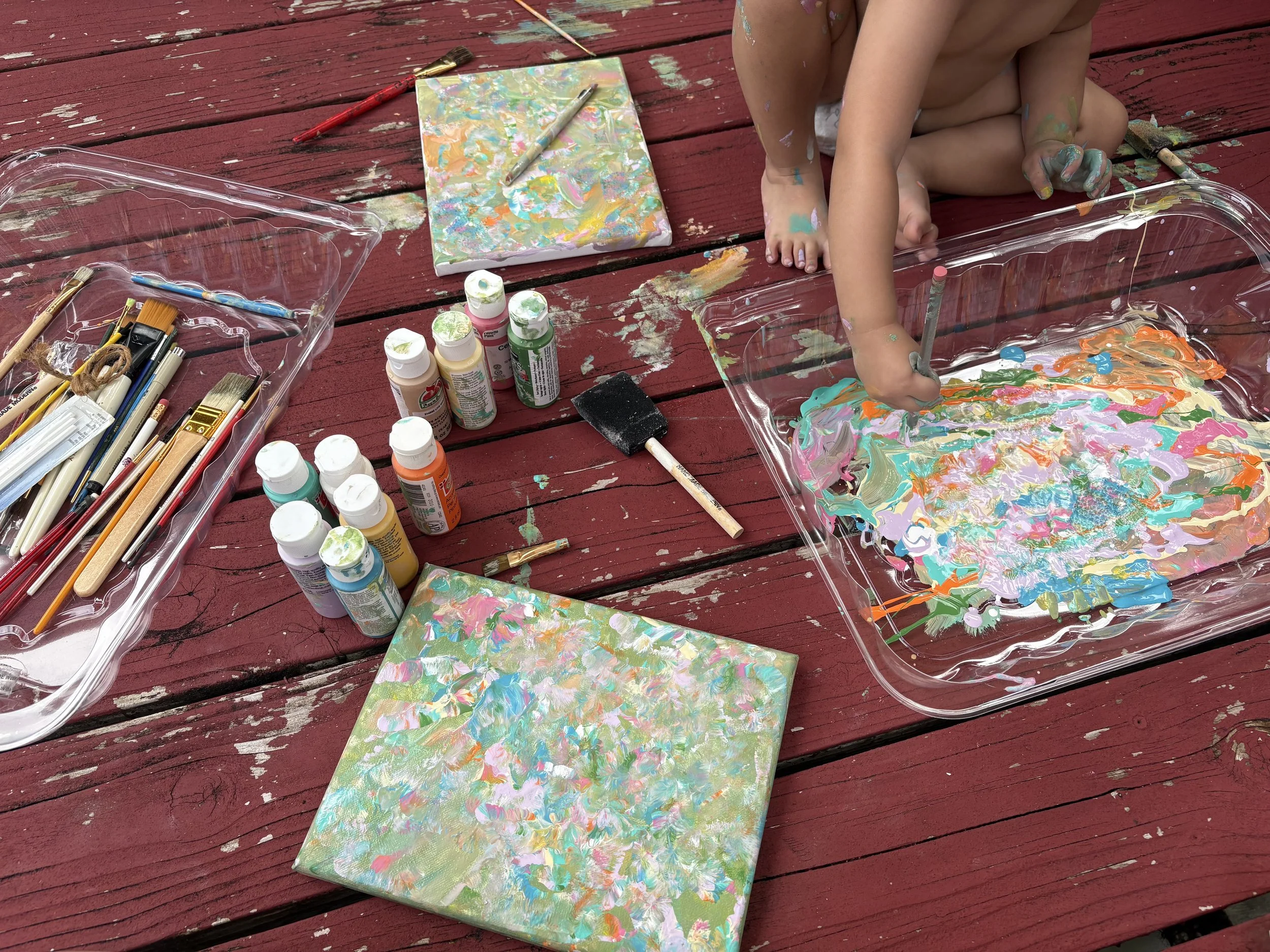 Child sitting on a red wooden deck, painting with colorful paints on a glass tray, surrounded by paint bottles, brushes, and blank canvases.