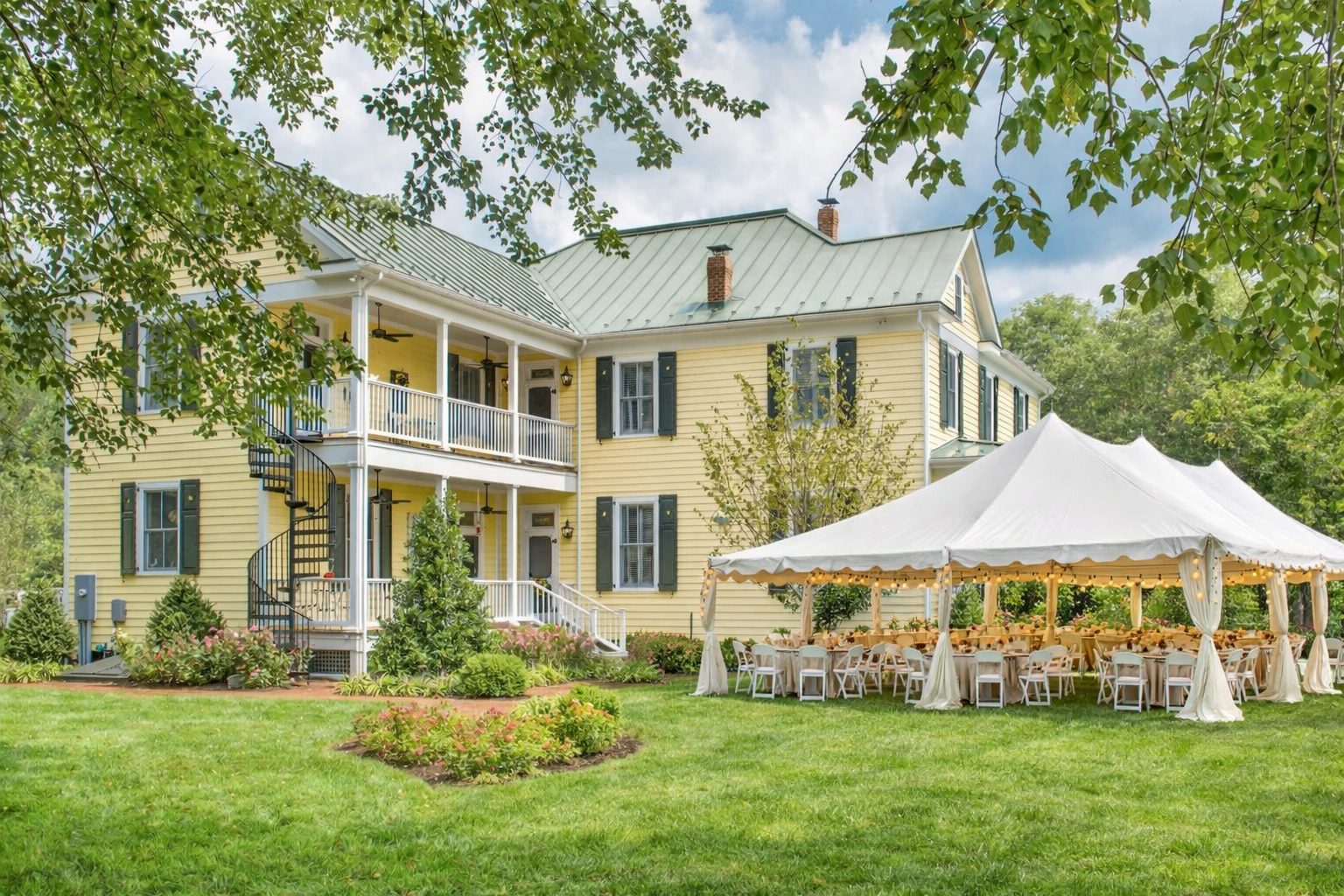 A yellow two-story house with green shutters and a green metal roof, surrounded by lush green trees and a well-kept lawn. A large white tent with string lights is set up in the yard, with tables and chairs inside for an outdoor event.