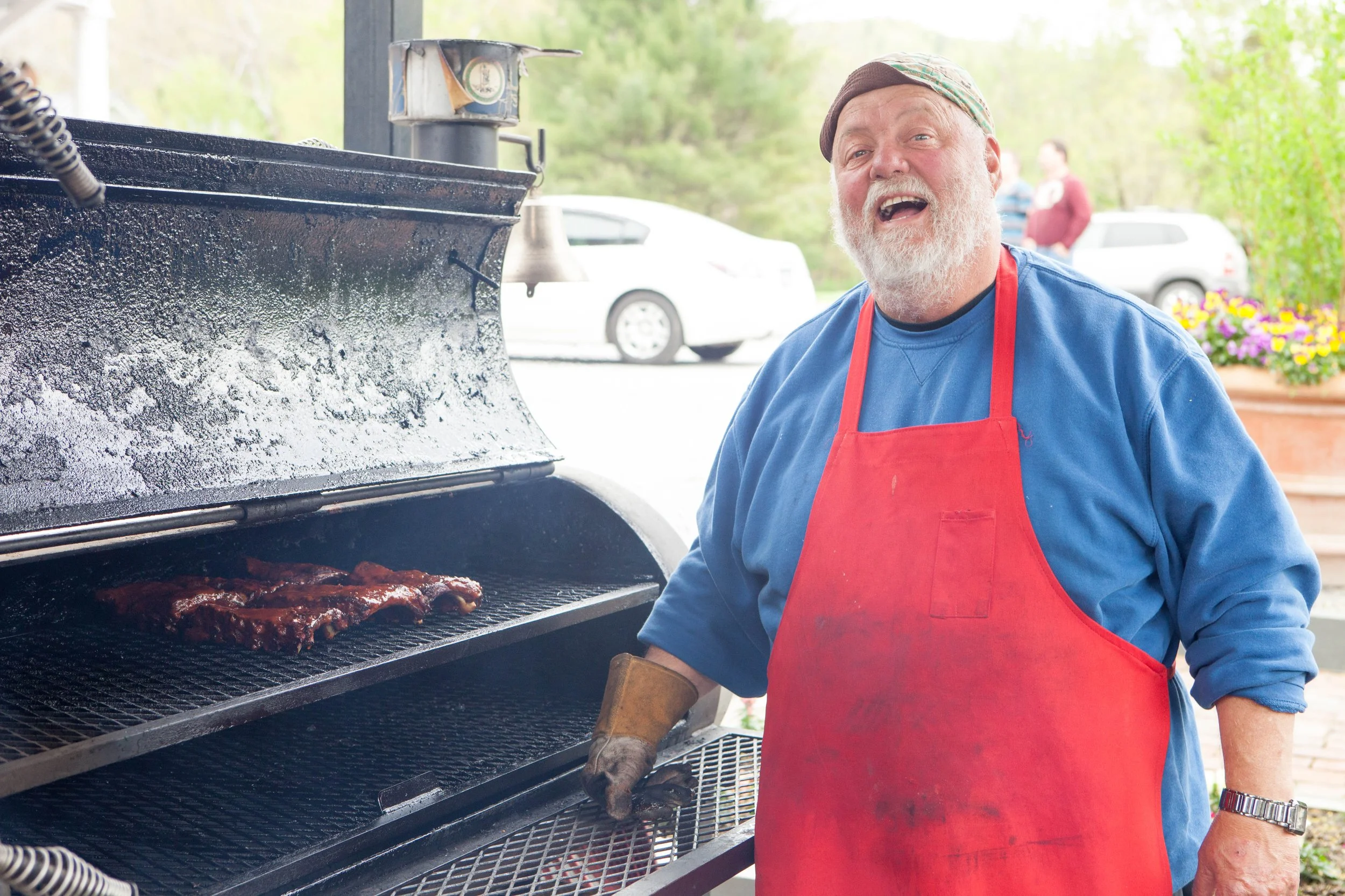 A man wearing a red apron and a blue shirt is standing next to a large outdoor grill with ribs cooking on it, smiling and talking.