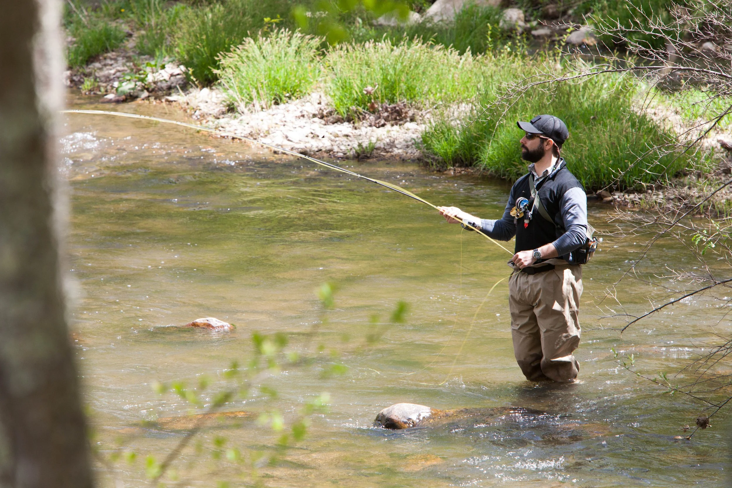 A man standing in a shallow river, fishing with a rod and reel, surrounded by trees and green bushes.