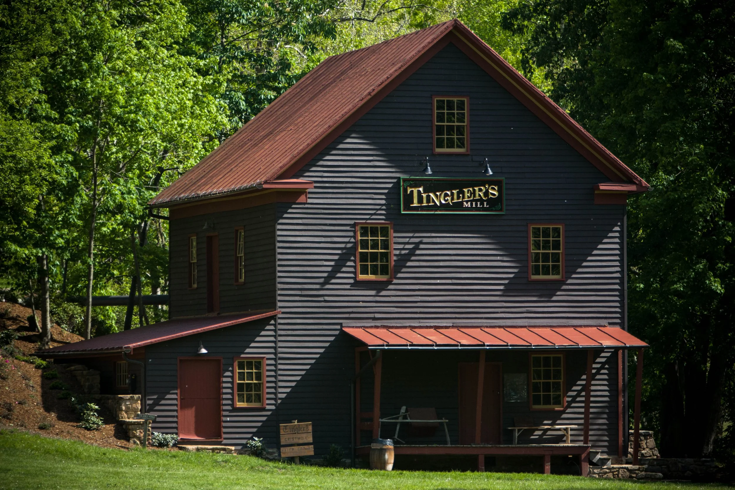 An old gray wooden building with a red metal roof, labeled "Tingler's Mill," surrounded by green trees.