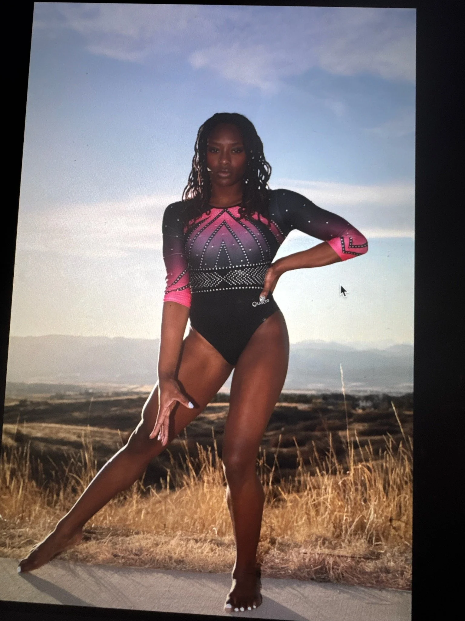 A woman posing in a black, pink, and purple athletic bodysuit with rhinestone embellishments, standing outdoors on a paved road with a field and mountains in the background during sunset or dawn.