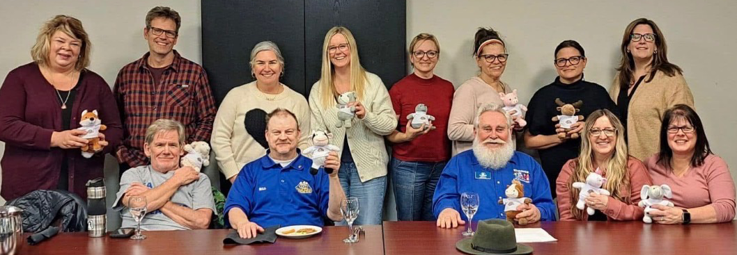 Group of people in an office setting holding plush toy animals, sitting and standing around a conference table with glasses and plates.