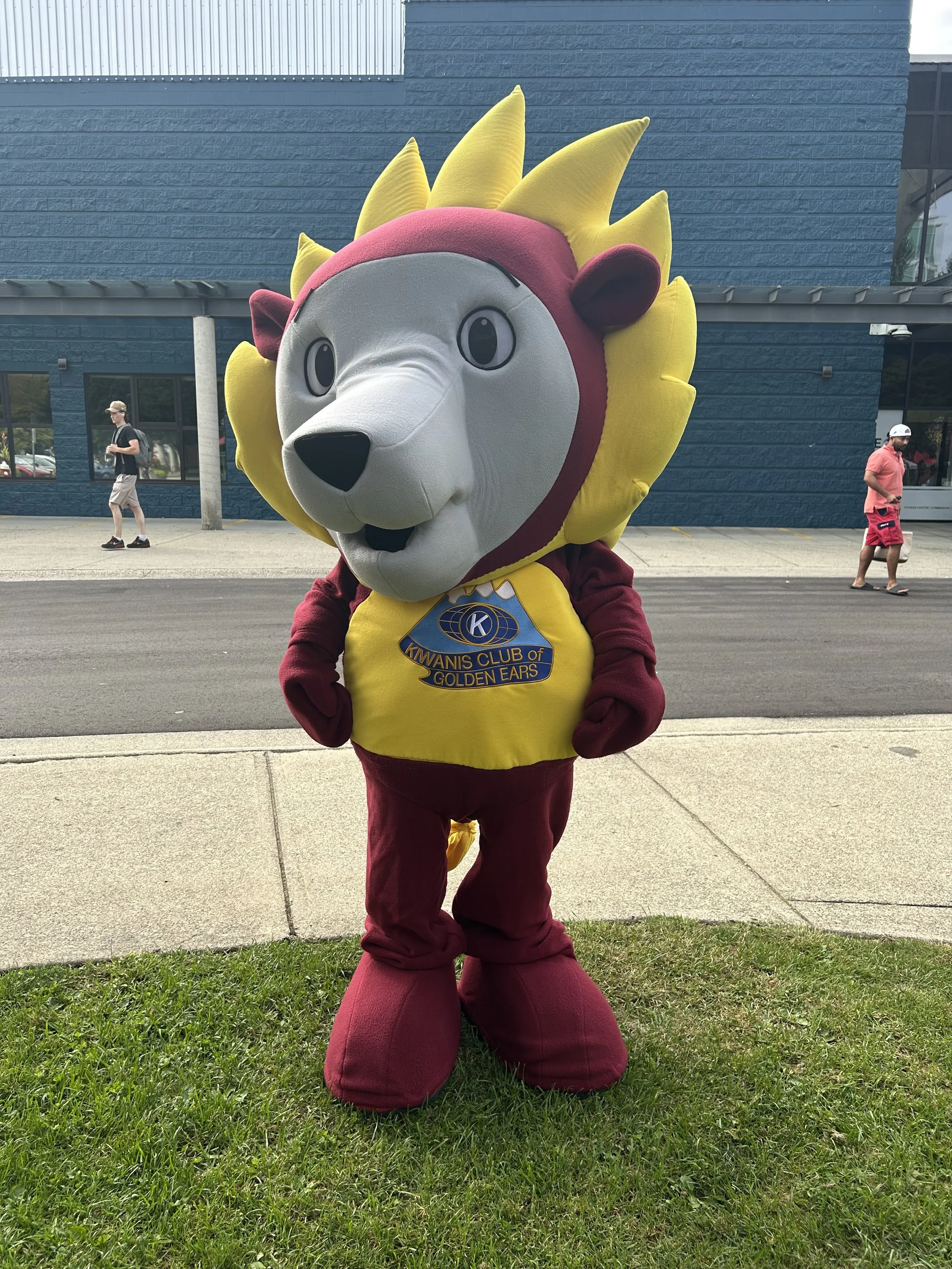A person in a lion mascot costume wearing a yellow shirt with the Kfans Club logo, standing on grass outside a blue brick building.