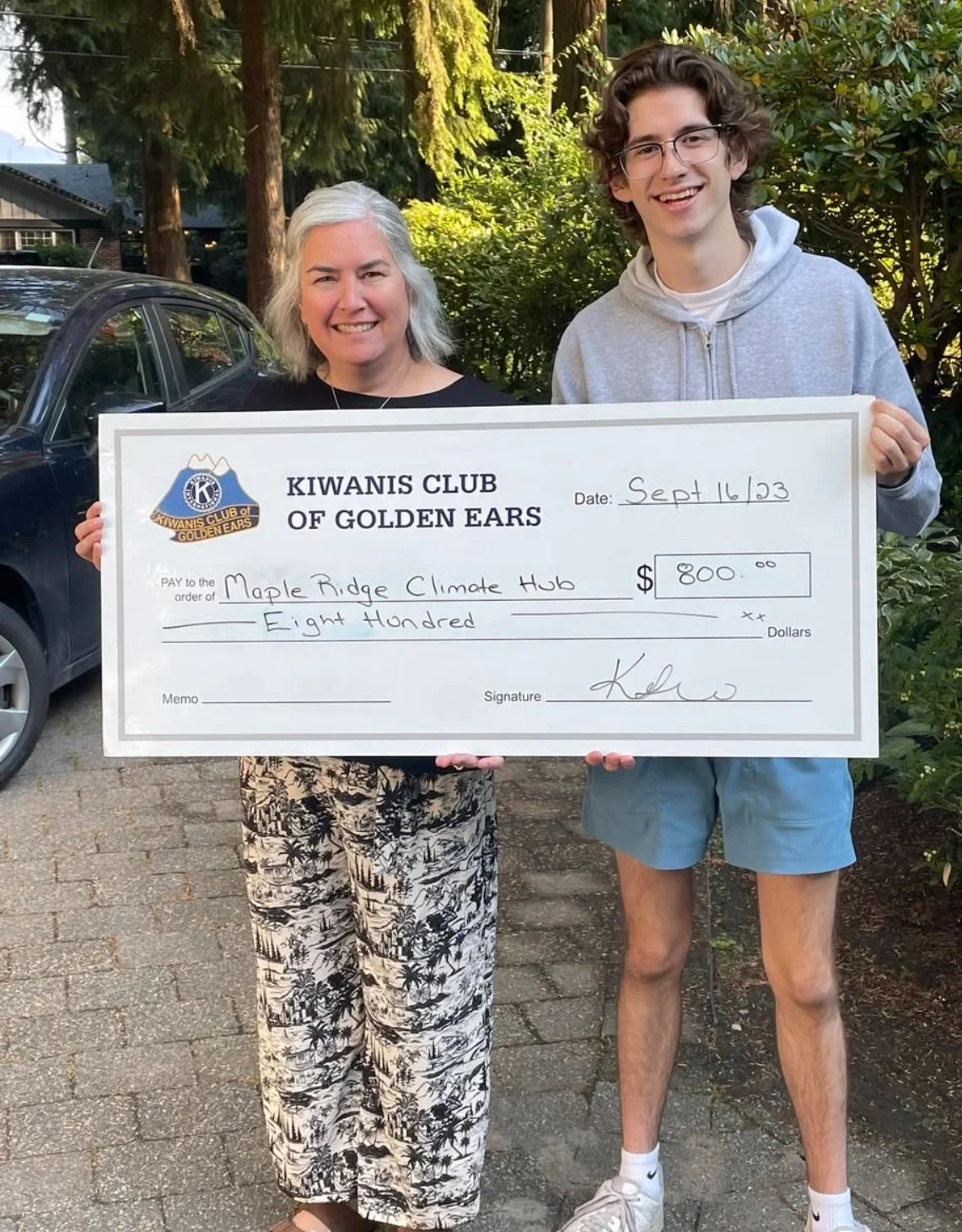 Two people holding a large check from the Kiwanis Club of Golden Ears, dated September 16, 2023, made out to Maple Ridge Climate Hub for $800. The woman on the left has short gray hair and is wearing a black top and patterned pants. The young man on the right has curly brown hair, glasses, a gray hoodie, and blue shorts. They are standing outdoors with greenery and parked cars in the background.