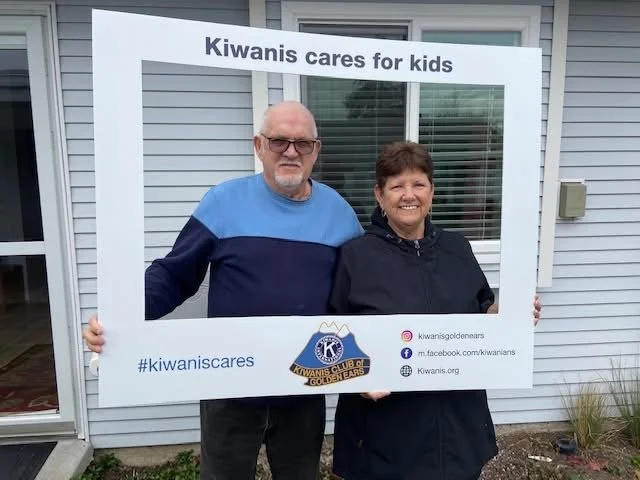 Two people holding a large frame with a banner that says "Kiwanis cares for kids" in front of a house with white siding and a window.