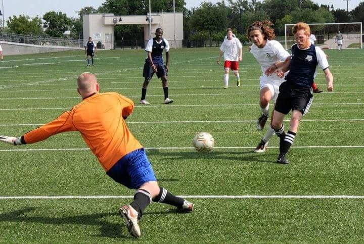Youth soccer game with players chasing the ball and goalkeeper trying to block shot on goal, on a grassy field with a goal post and trees in the background.