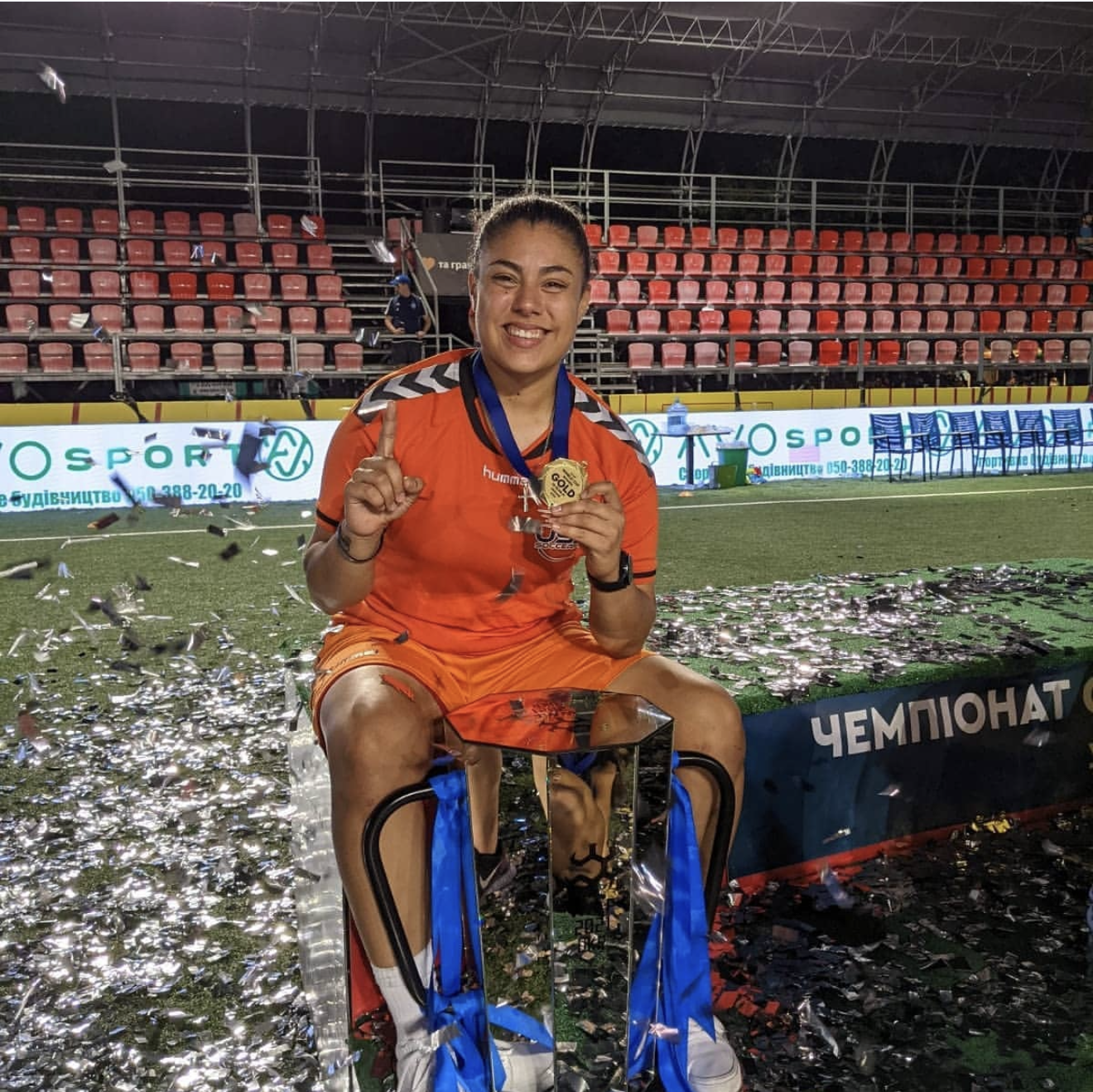 A female soccer player in an orange uniform sitting on a chair on the soccer field, smiling, holding a gold medal, with a blue ribbon around her neck. Confetti is falling, and a banner with Cyrillic text is visible in the background.