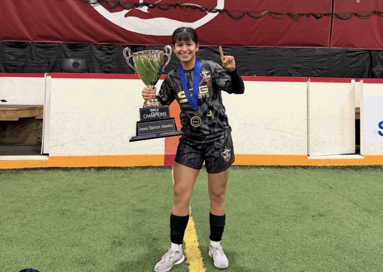 A young female athlete in black sports attire with a medal around her neck, holding a large trophy, standing on an indoor sports field, and smiling at the camera.