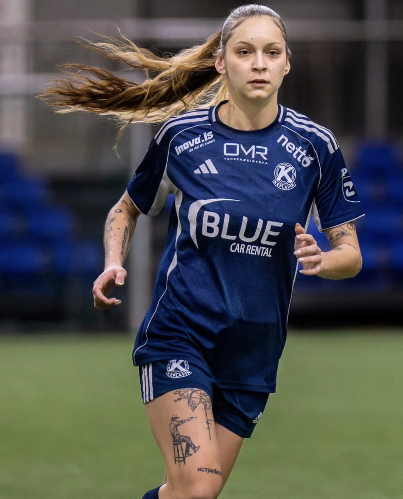 A female soccer player with long hair running on the field wearing a blue jersey with various sponsor logos, including Blue Car Rental, and having tattoos on her left arm and right thigh.