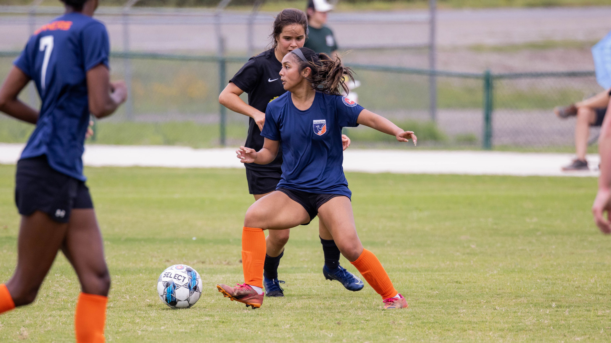 Women playing soccer on a grassy field with a ball, with a fence and spectators in the background.