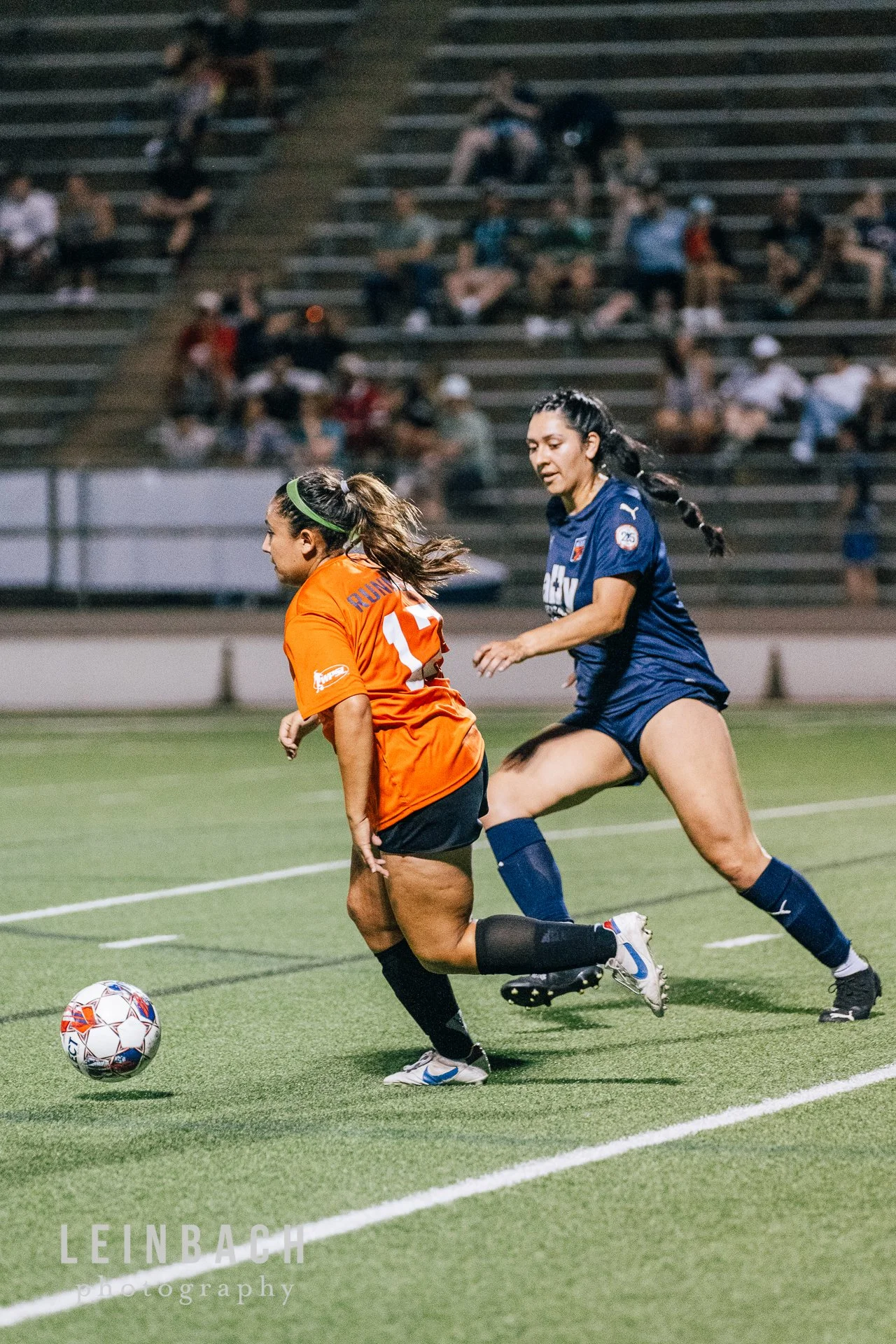 Two female soccer players competing for the ball on a green field during a match at night. One player is wearing an orange jersey and black shorts, the other is in a blue jersey and shorts. Spectators are seated on bleachers in the background.