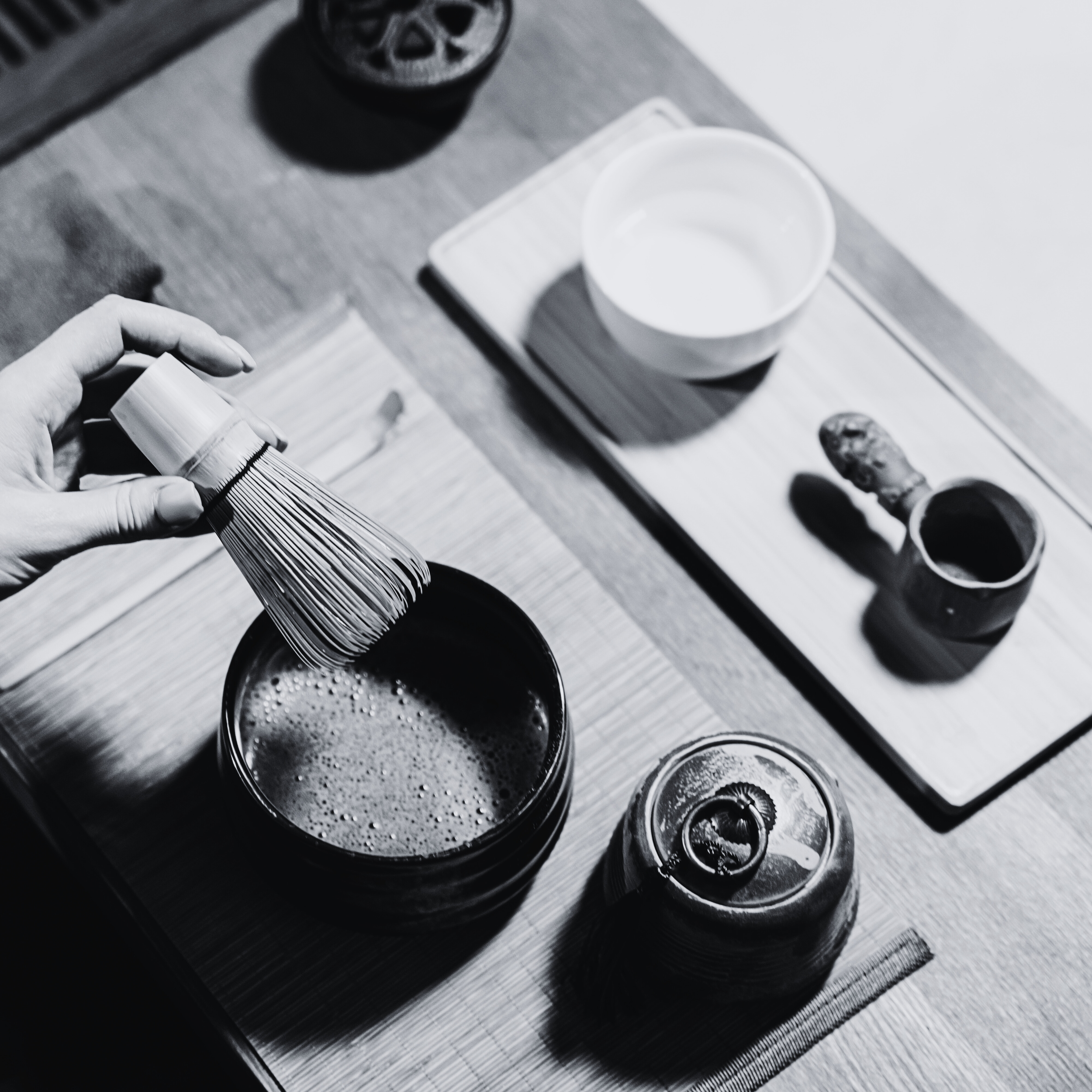 A person holding a tea whisk over a small black bowl of frothy tea, with various tea accessories on the table including a jar with a stir stick, a white cup, and a tray with a teapot, cup, and a small container of tea powder.