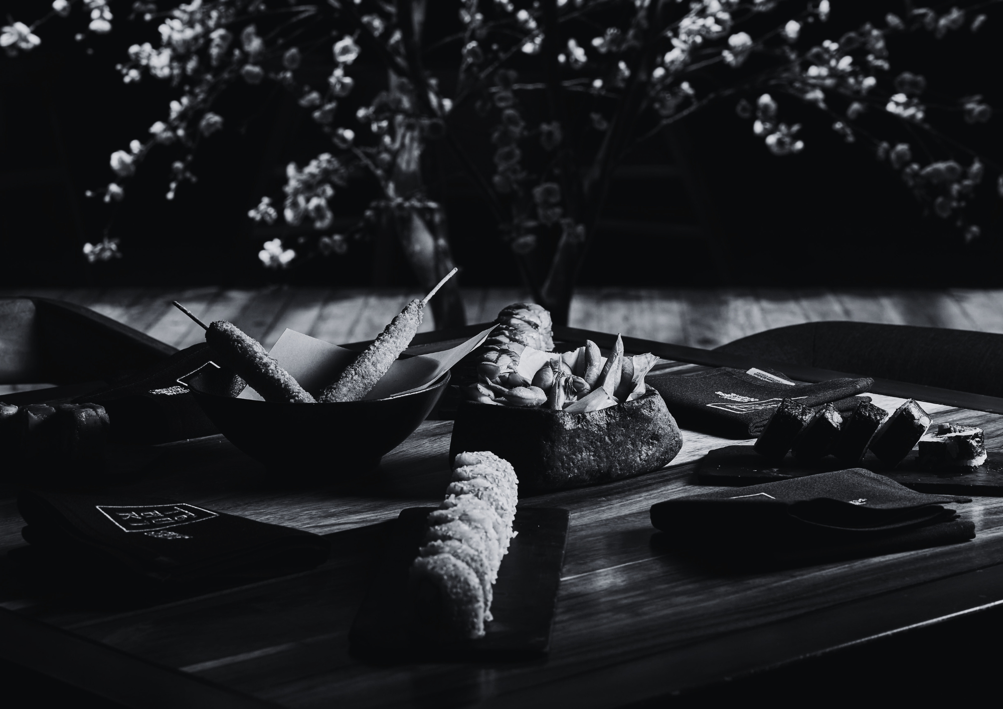 A black and white photo of a wooden table with various food items, including skewered snacks and wrapped treats, with a backdrop of hanging flowers.