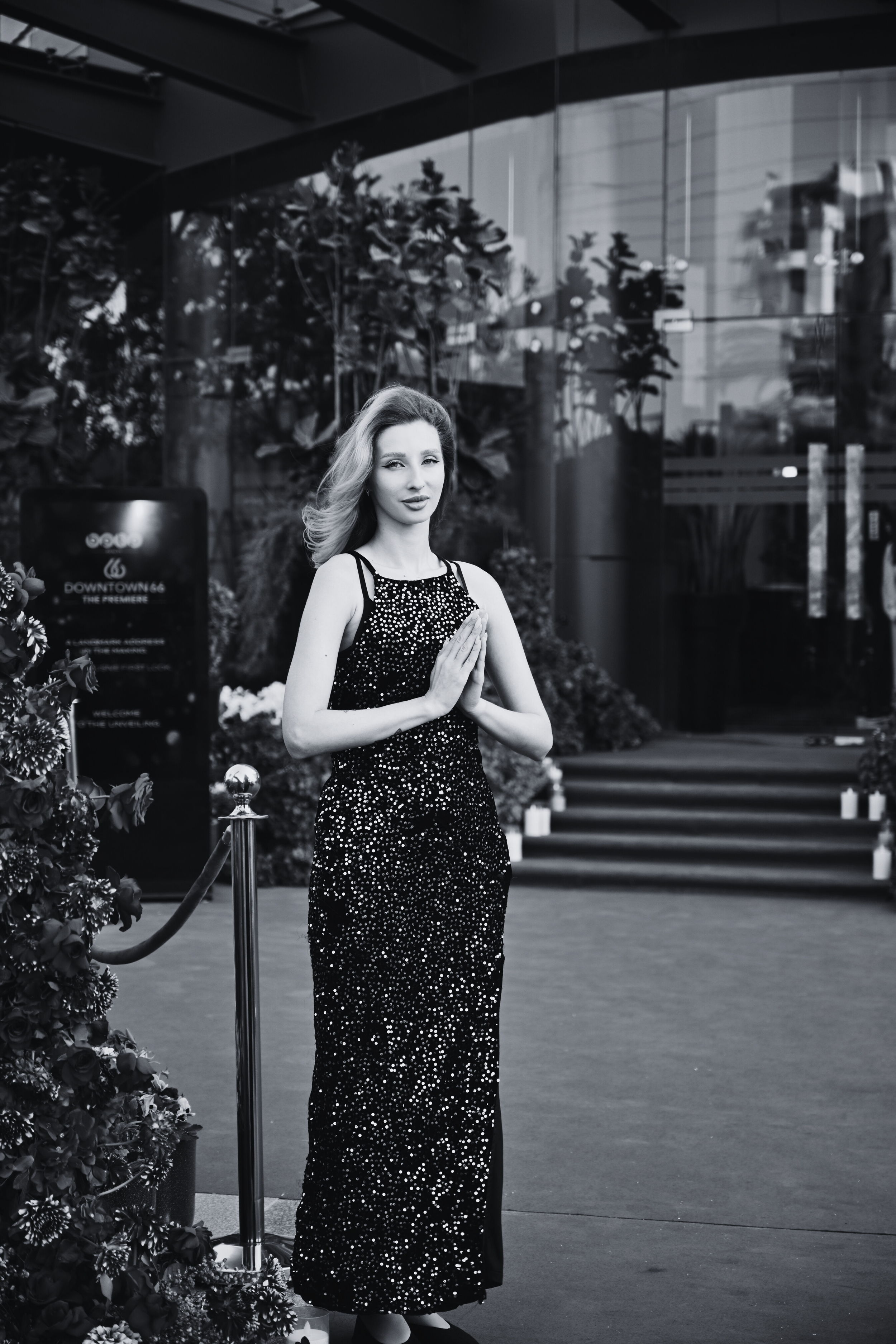 A woman in a sparkly black evening gown stands with her hands pressed in a prayer position outside a building with stairs and glass walls.