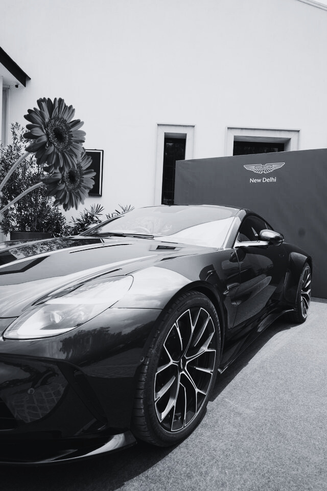 A black sports car parked and a banner with the Aston Martin logo and 'New Delhi' written on it.