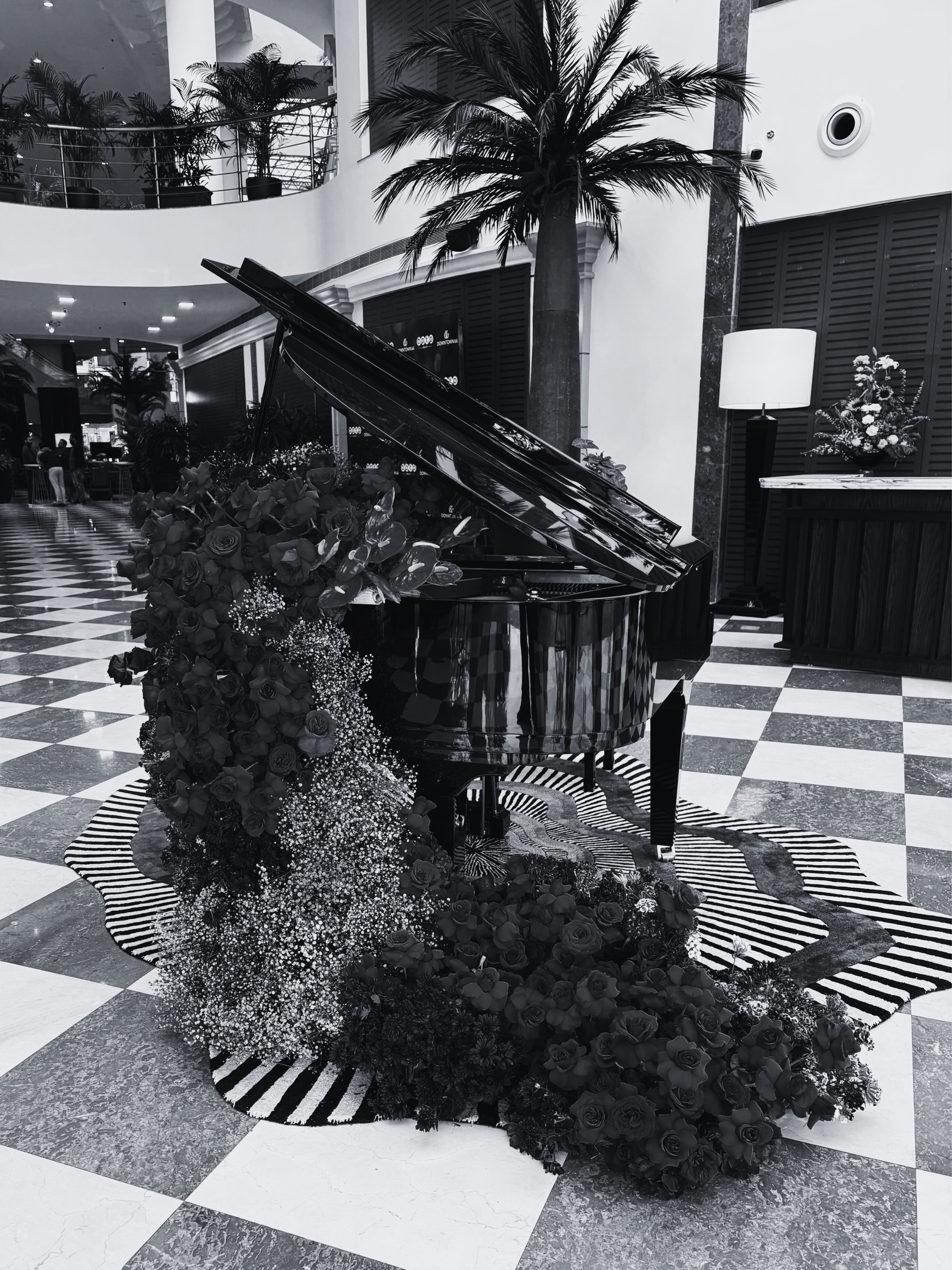 A black grand piano decorated with flowers and baby's breath, placed on a patterned rug in a spacious indoor area with tropical plants and checkered flooring.