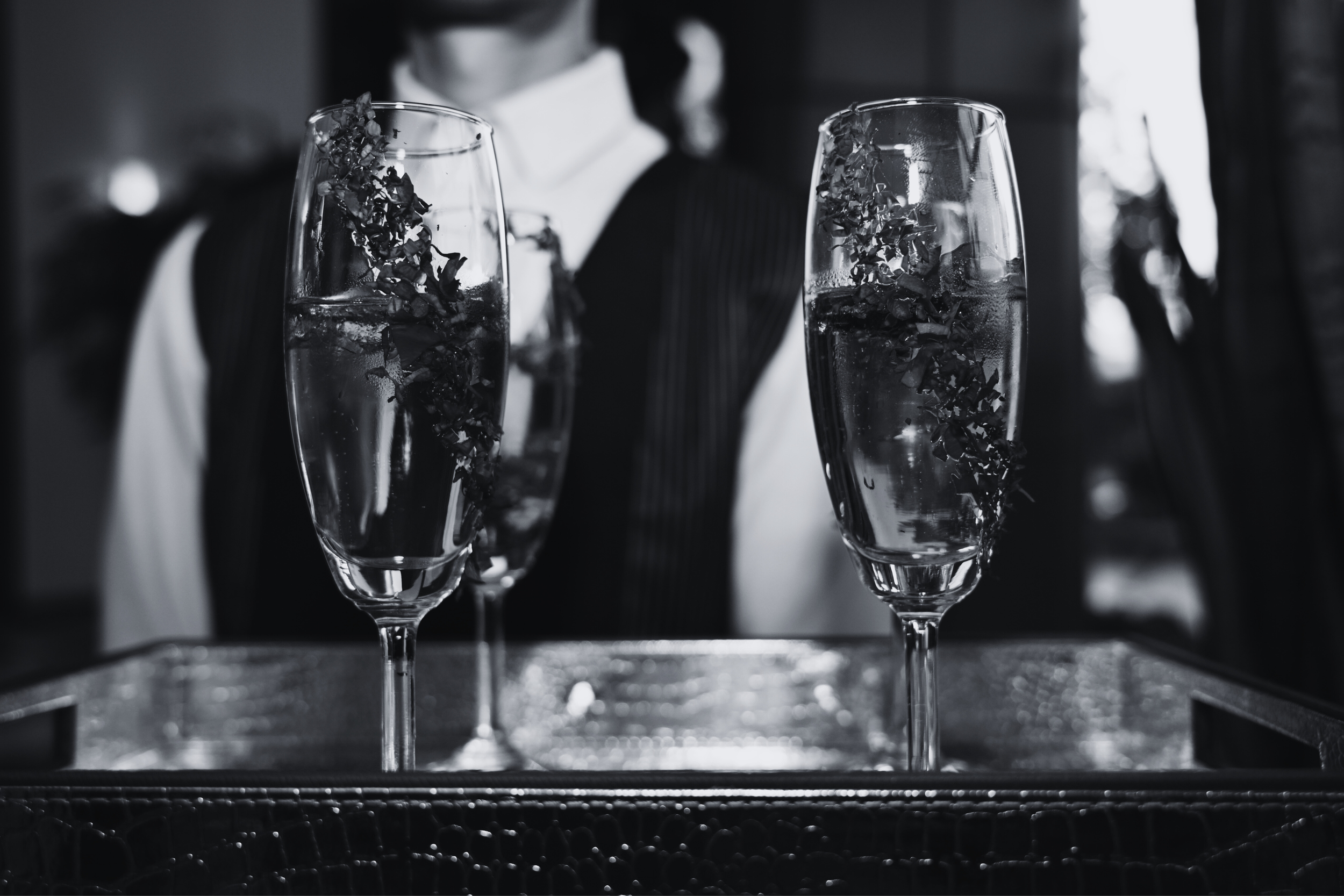 Two champagne glasses with ice and garnish, placed on a tray, with a person in formal attire in the background.
