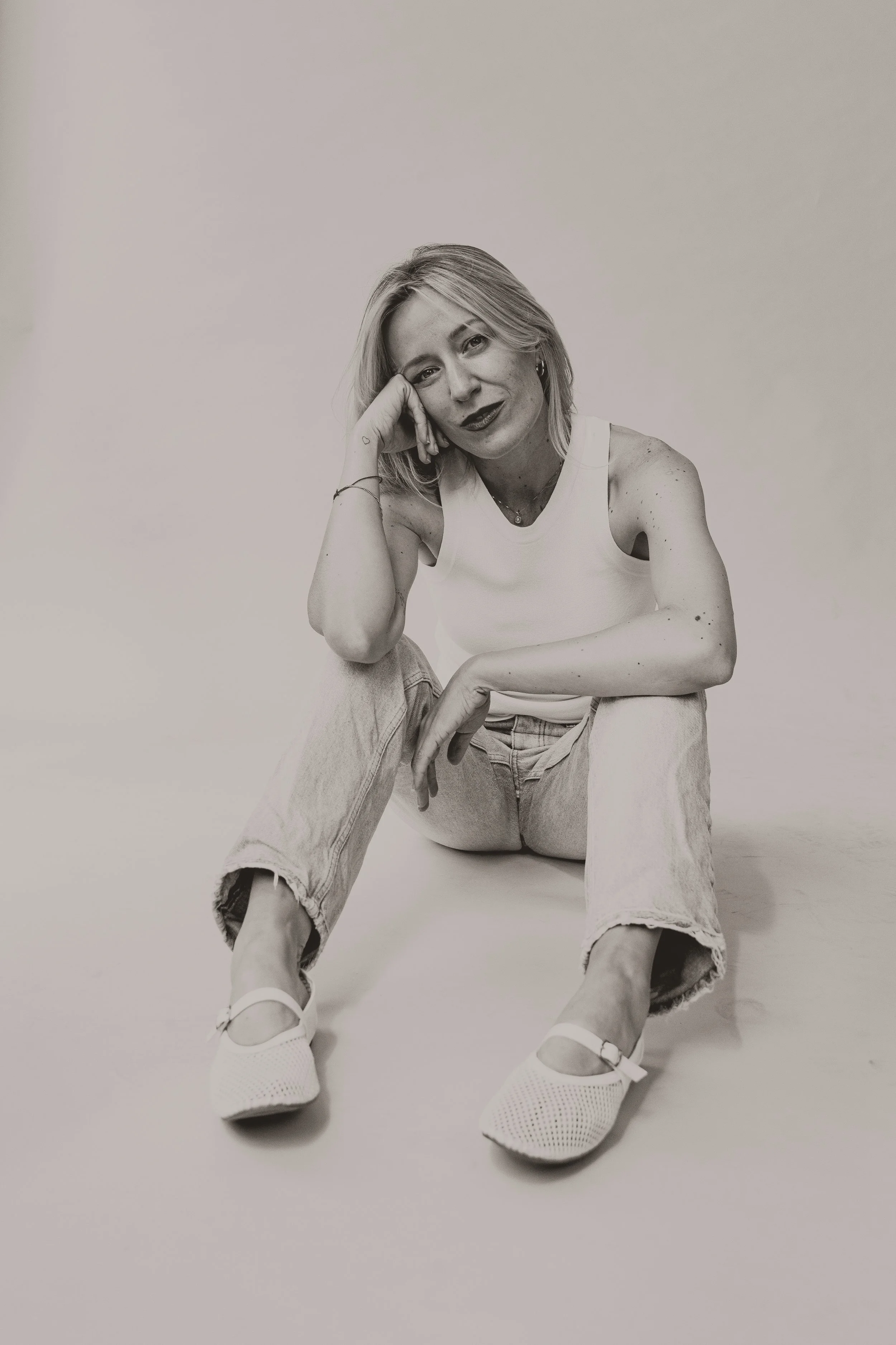 Black and white photo of a woman sitting on the floor, wearing a sleeveless top, jeans, and sneakers, with a relaxed pose and hand resting on her chin.