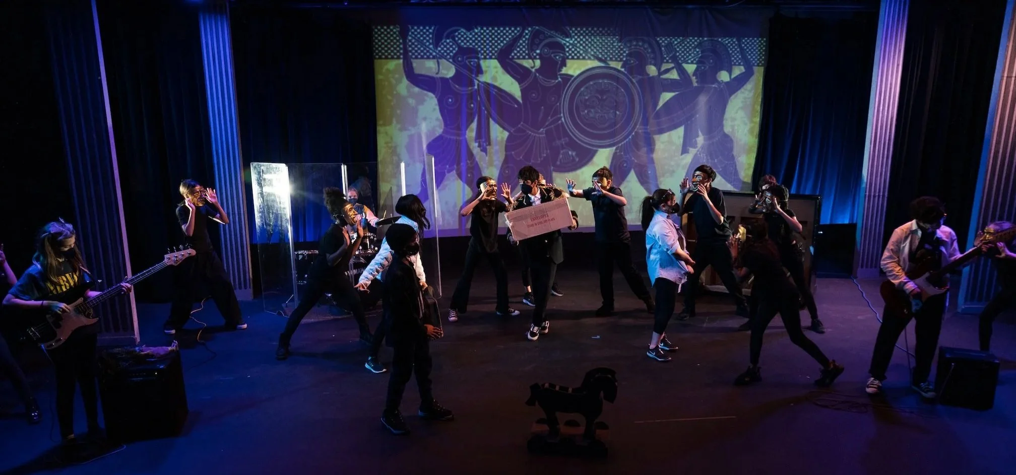 A group of people wearing face masks are performing on a stage with blue and purple lighting. Some are playing guitars, others are dancing, and a large screen in the background displays a graphic of ancient warriors with shields and spears.