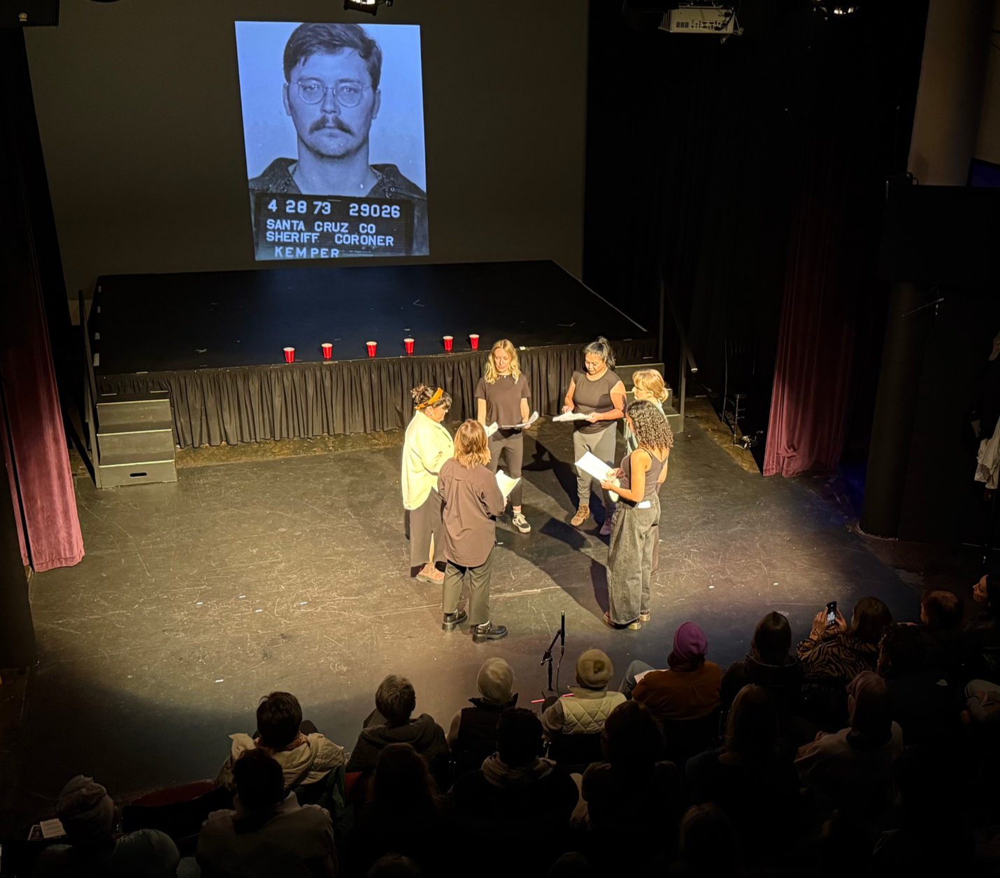 A stage with six women holding scripts, engaging in a rehearsal or performance, with an audience watching from the seats. On the wall above the stage, a large projected image of a mugshot of a man with a mustache and glasses, with text indicating 'Sa