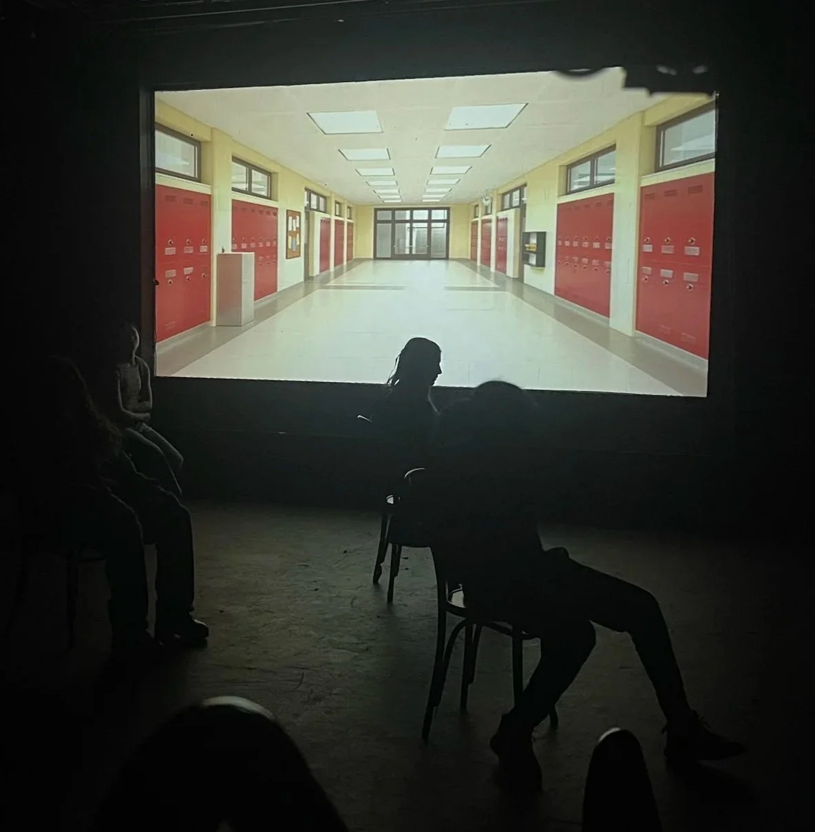 People watching a film in a dark room with a school hallway on the screen, showing red lockers, windows, and a door at the end.