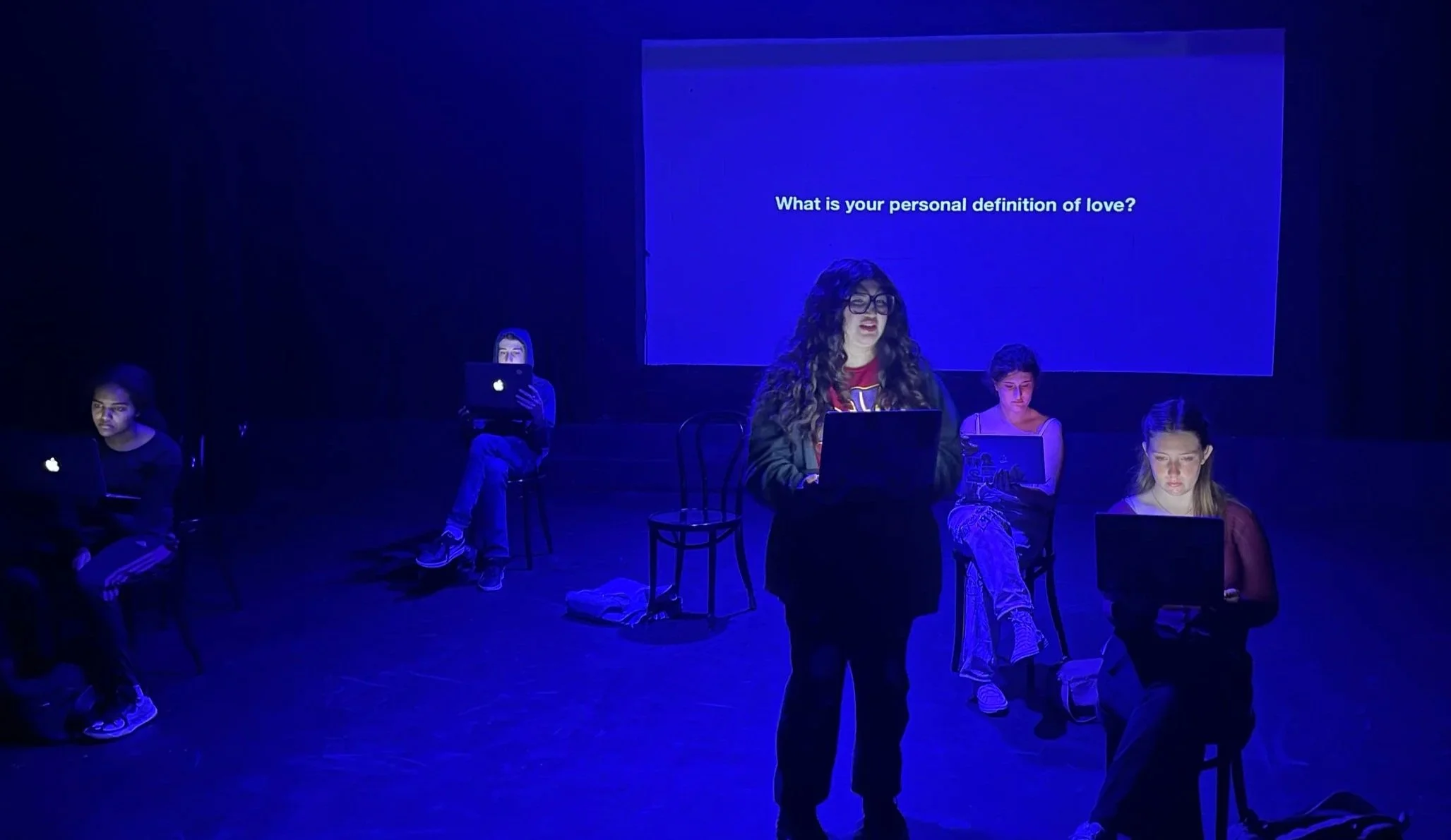 A woman standing in front of a group of women sitting on chairs in a dark room with a blue backdrop and a screen that asks, 'What is your personal definition of love?'