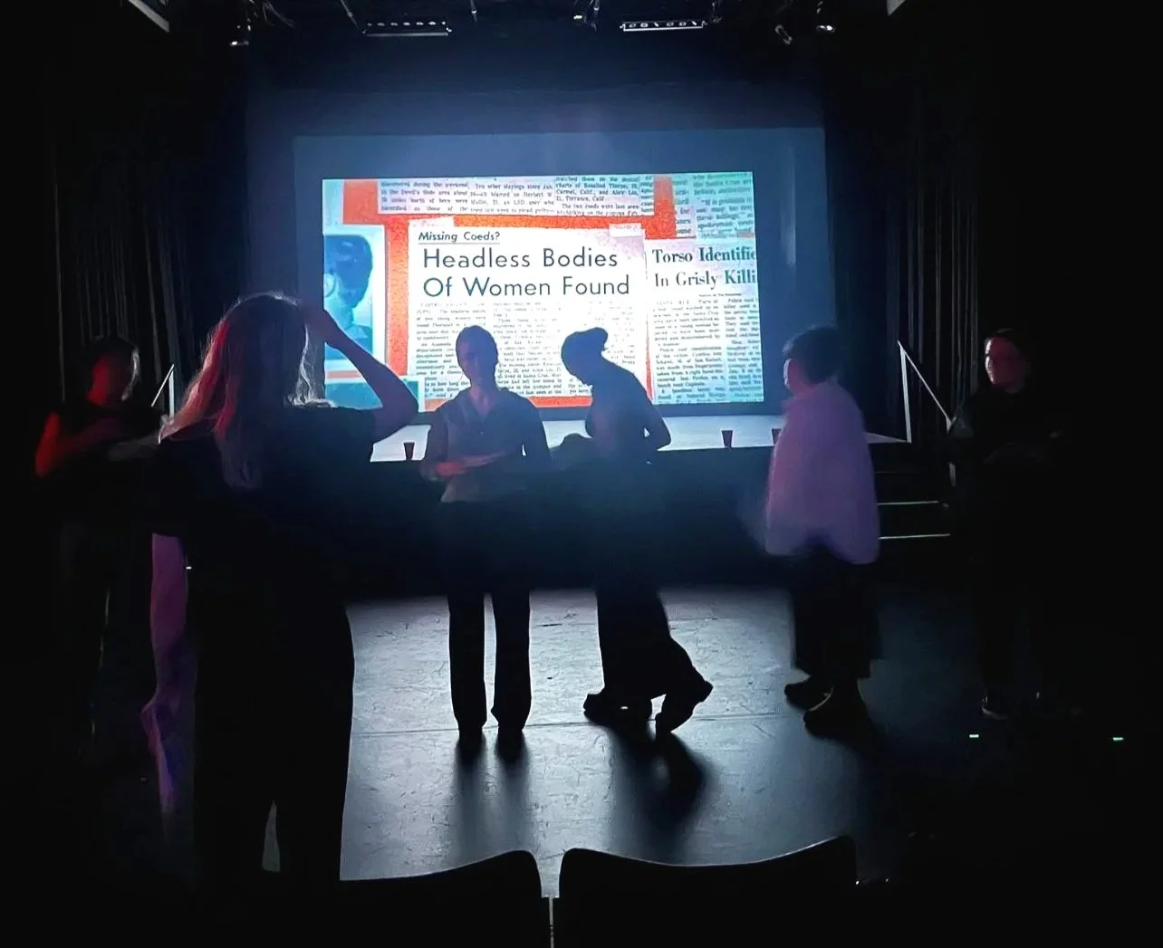 A group of six people, mostly women, standing and talking in front of a large screen displaying a news headline about headless bodies of women found. The scene is dimly lit, with the bright screen providing backlighting.