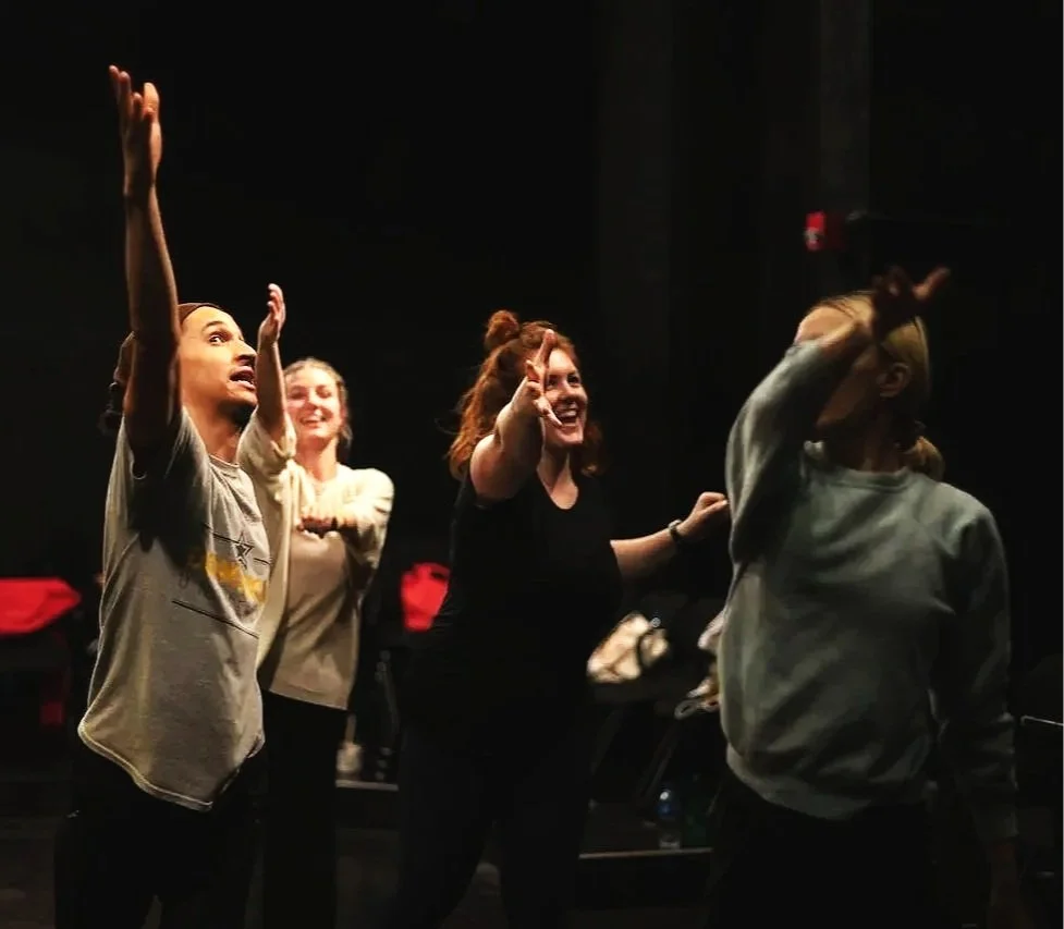 Four women in a dark room, two of them with arms raised and smiling, engaging with someone or something out of frame.