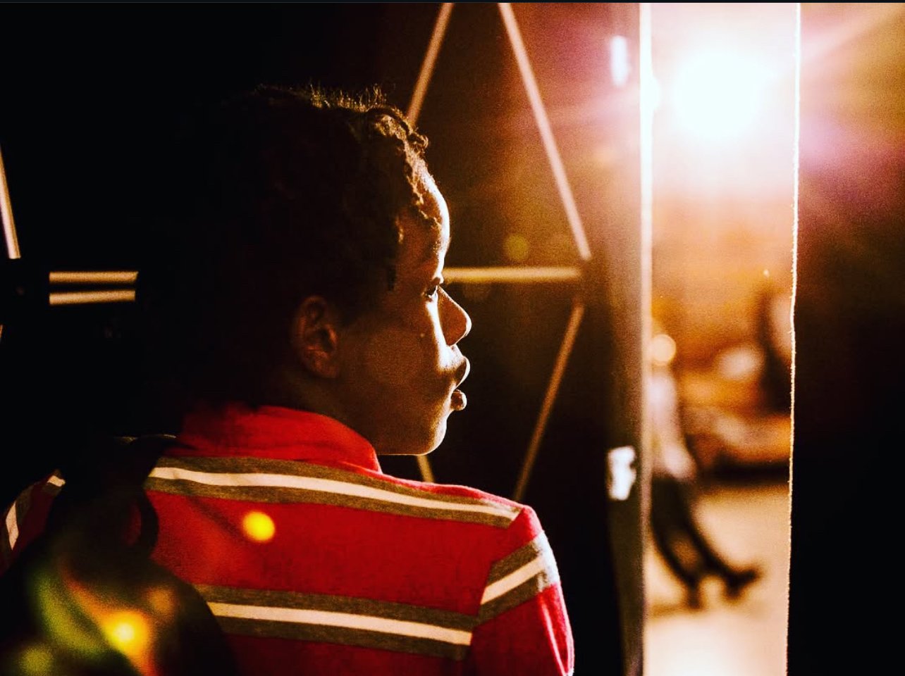 A woman with natural hair in profile, illuminated by warm light, sitting in a studio or stage setting.
